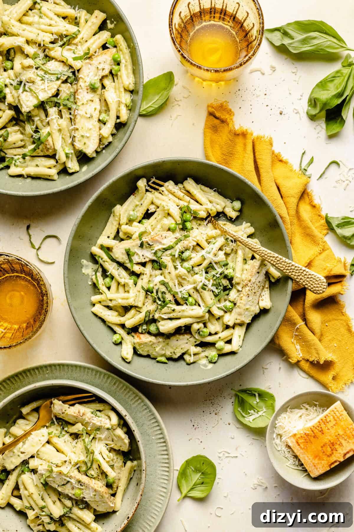 A top down view of a white countertop with three sage-green dishes of Creamy Pesto Chicken Pasta. There are also two yellow glasses of water, various basil leaves, mustard colored napkins, and a dish of a block of parmesan cheese.
