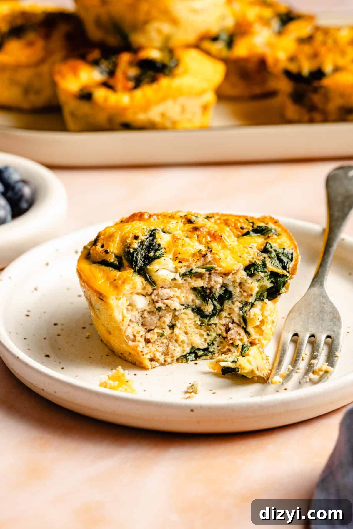 A finished Sausage and Egg Mini Frittata on a white plate with a silver fork. A bowl of blueberries is visible in the background.
