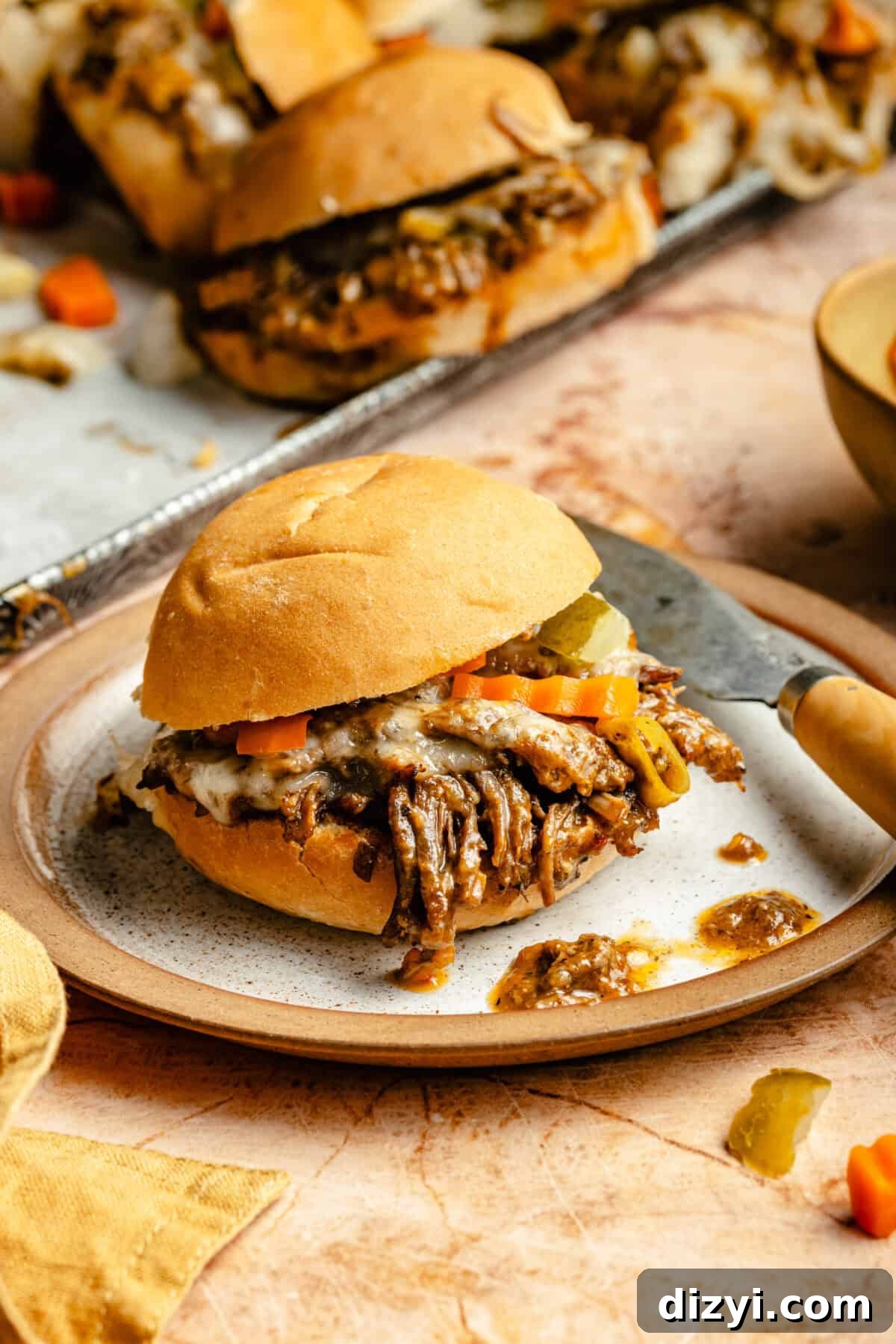 Slow Cooker Italian Shredded Beef Sliders Recipe 4 A close up shot of a Slow Cooker Italian Shredded Beef Slider on a plate on a terracotta colored countertop. A tray of finished Slow Cooker Italian Shredded Beef Sliders sits in the background.