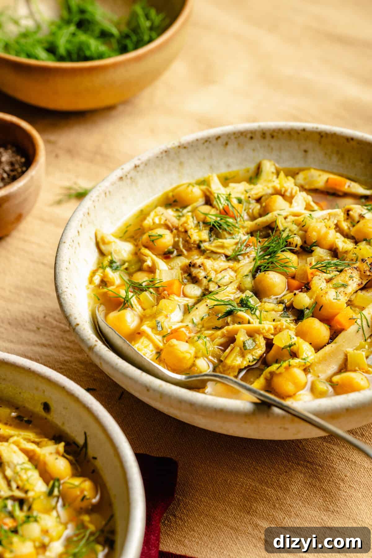 One-Pot Lemon Chicken and Chickpea Stew 2 A finished bowl of Lemon Chicken and Chickpea Stew in a cream colored dish, with a silver spoon. A bowl of herbs and a small bowl of spices are visible in the background. The dishes sit on a pale yellow napkin.