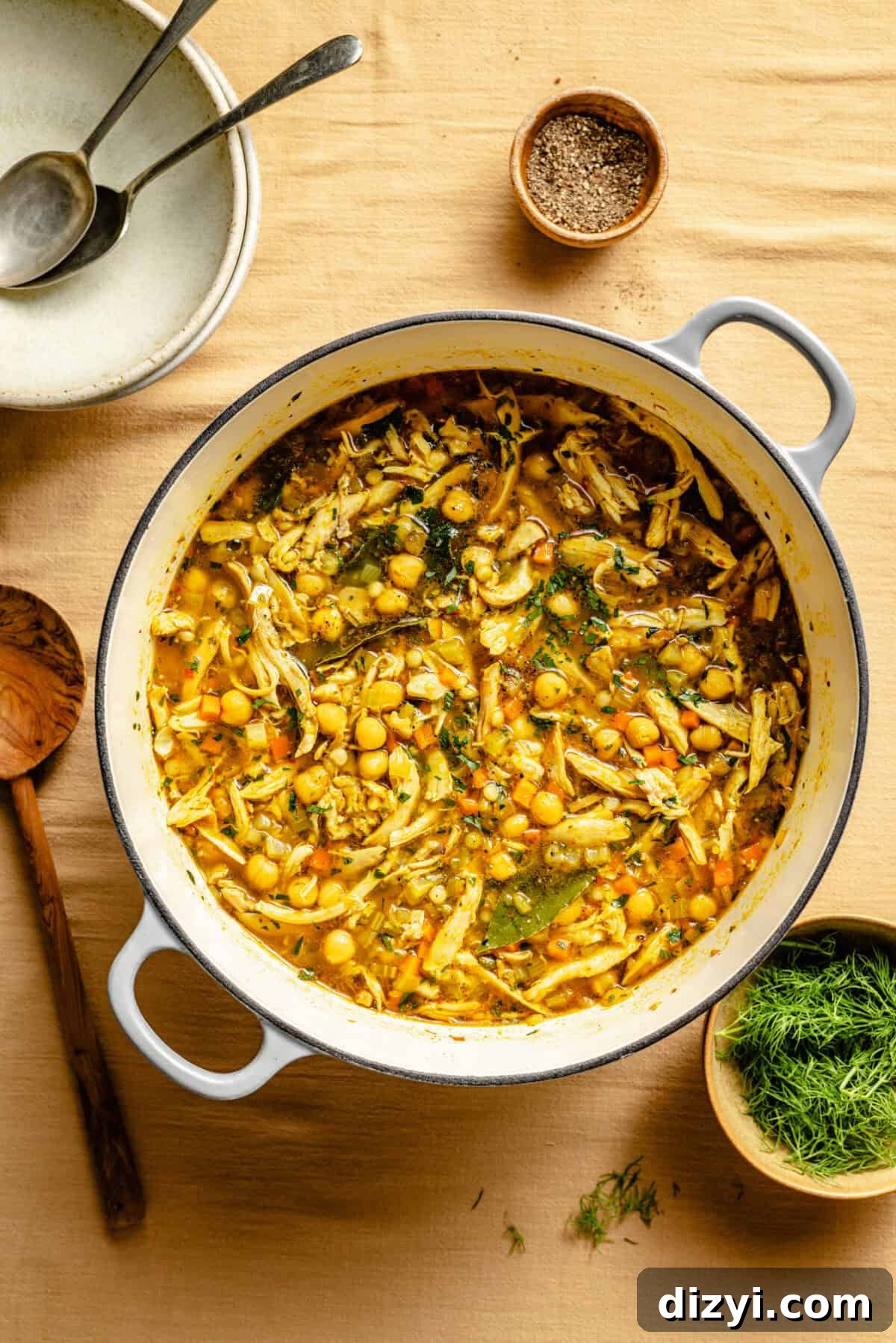 One-Pot Lemon Chicken and Chickpea Stew 5 The stockpot of Lemon Chicken and Chickpea Stew sitting on a yellow tablecloth. Small dishes of spices and herbs are visible. A wooden serving spoon sits next to the pot, along with two bowls and two spoons, waiting to be served.