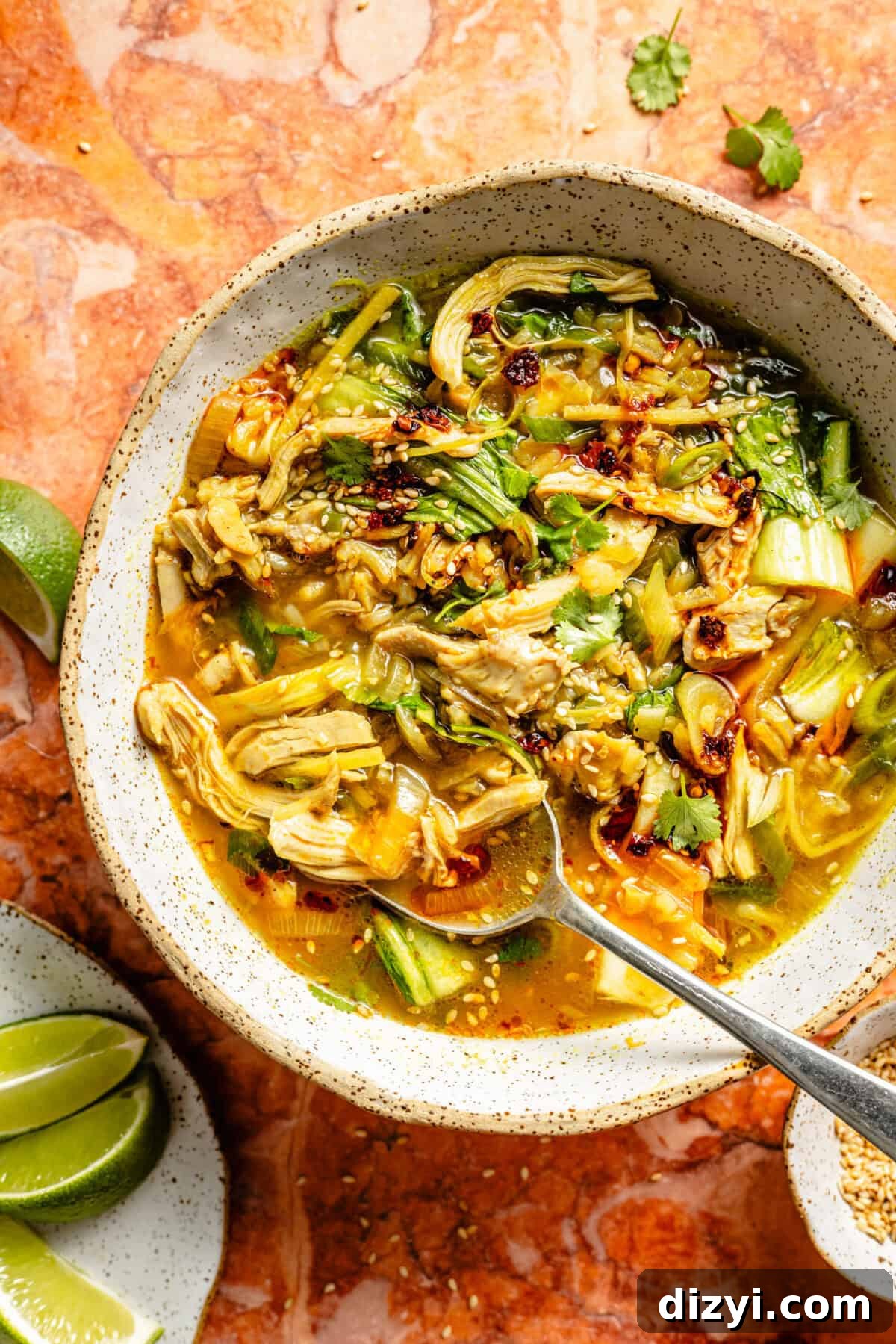 A finished bowl of Ginger-Scallion Chicken and Rice Soup in a speckled dish. There is a silver spoon, a small dish of limes, and a few sprigs of cilantro all atop an orange countertop.