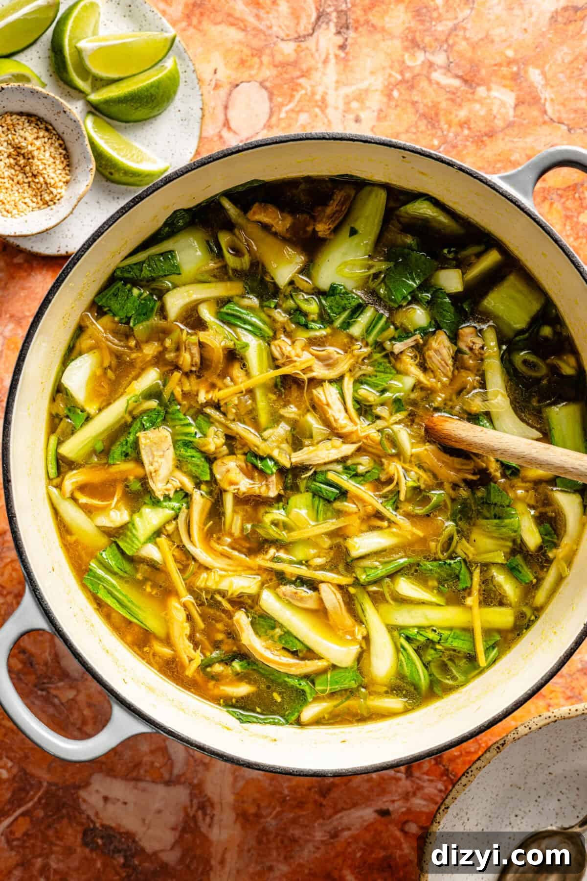 A stockpot of Ginger-Scallion Chicken and Rice Soup simmering on top of an orange countertop. A small white dish with spices and lime wedges is at the top of the image.
