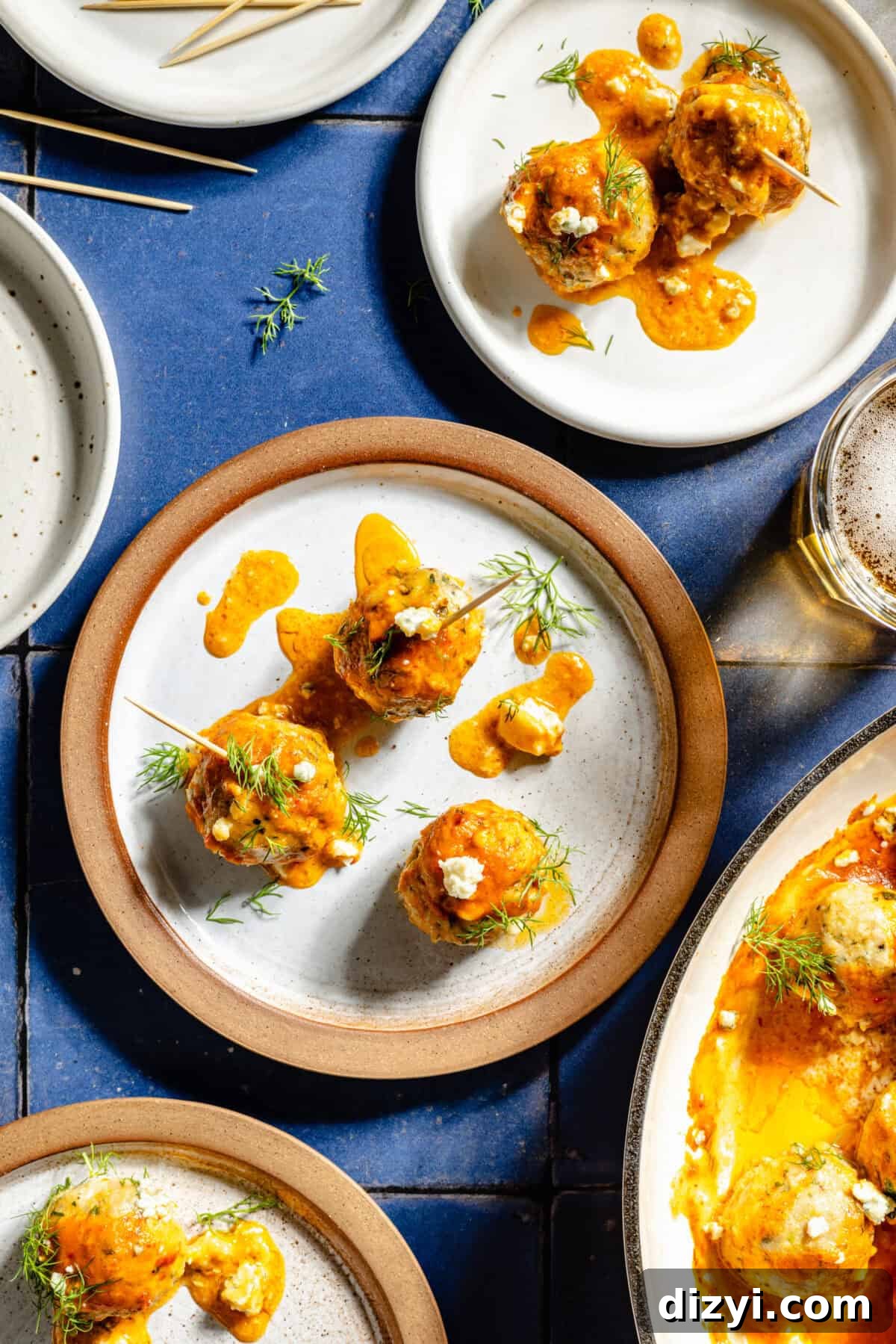 Plated Buffalo Chicken Meatballs on white and tan plates served with toothpicks. All of the dishes rest on a blue countertop.