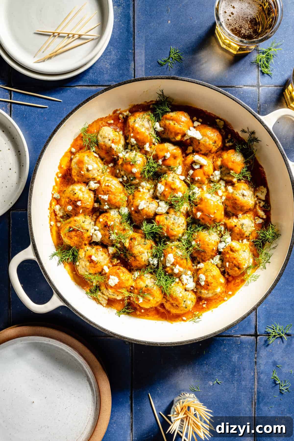 A top-down view of Buffalo Chicken Meatballs in a shallow stockpot on top of a blue tile countertop. A few dishes are just out of view, as well as a small dish of toothpicks for serving.