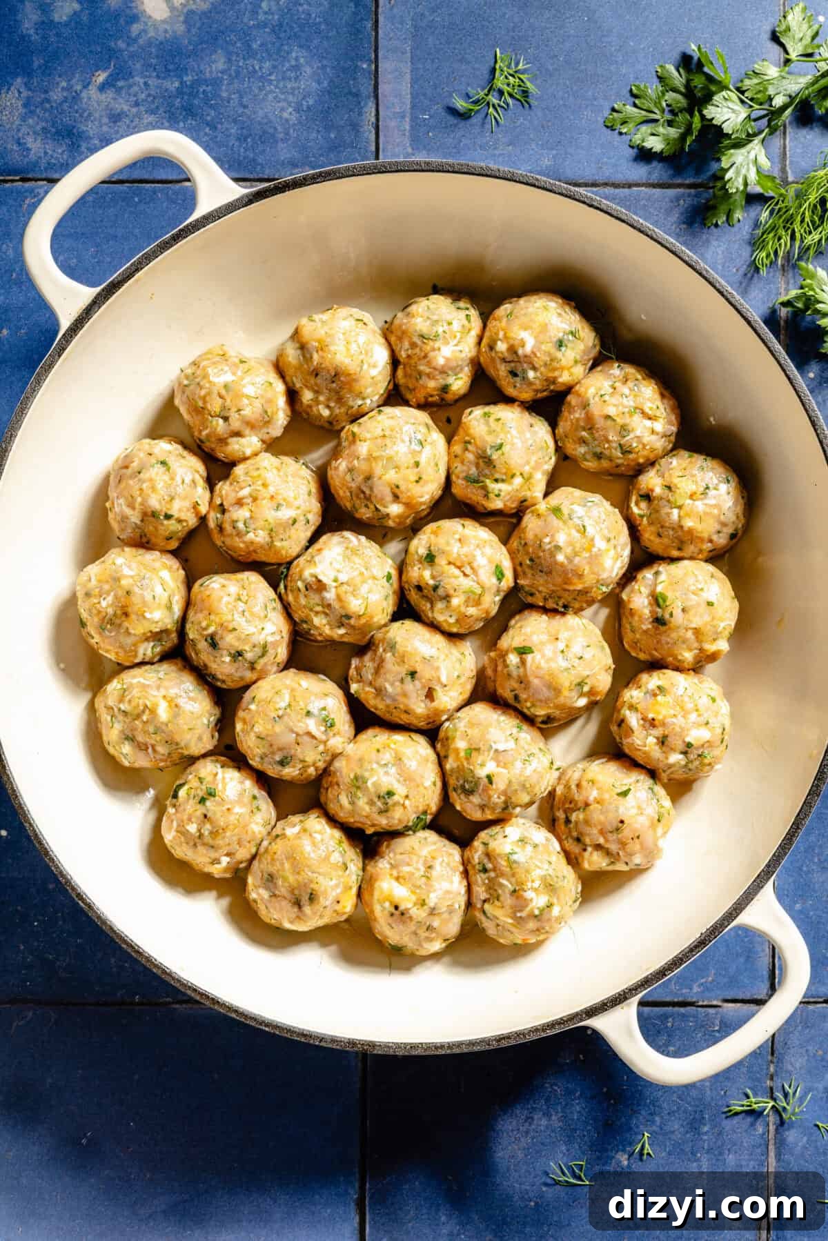 A shallow, white stockpot of Buffalo Chicken Meatballs ready to be cooked. The pot sits on top of a blue tile countertop, and there are a few sprigs of fresh herbs to the side.