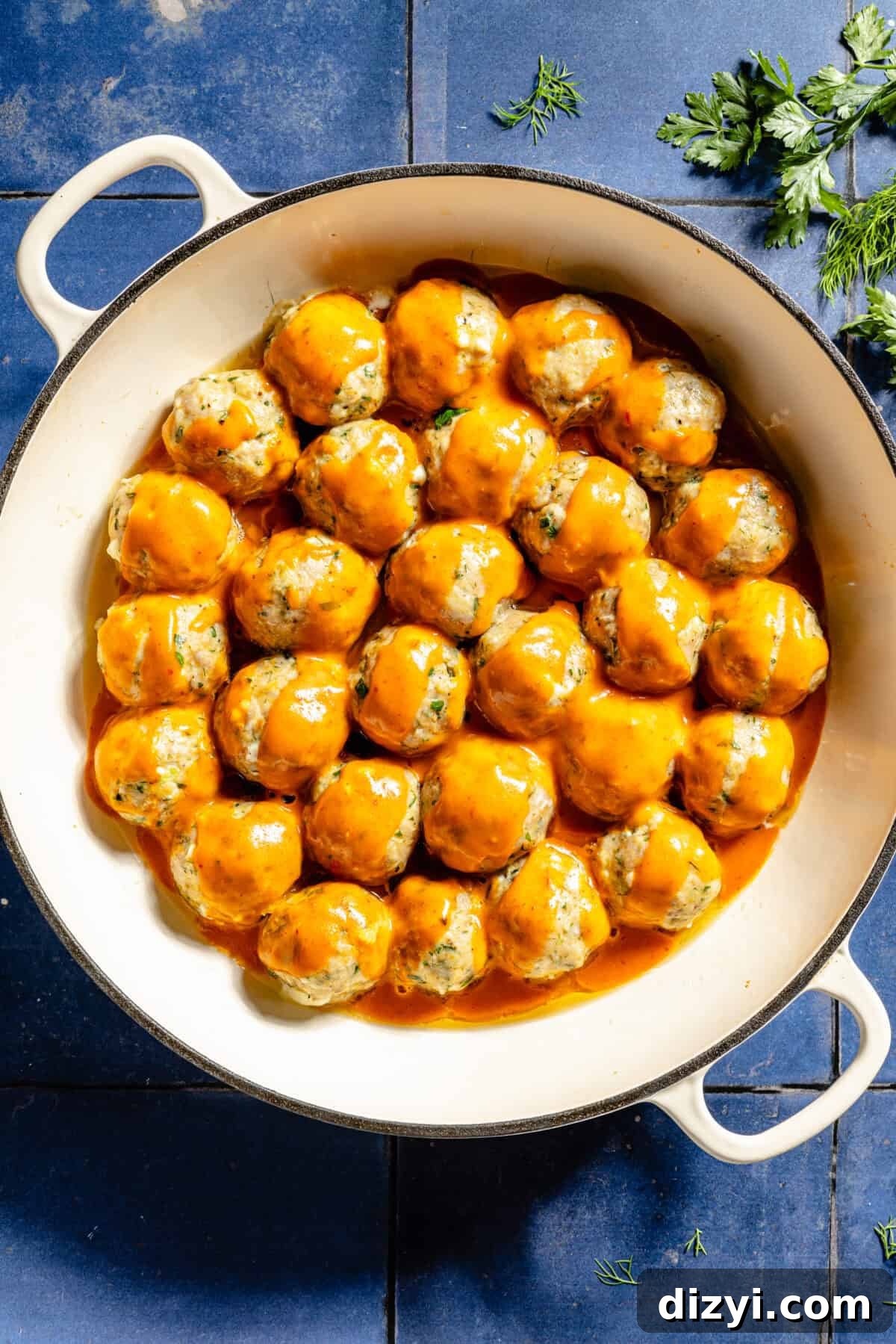 The Buffalo Chicken Meatballs with the sauce added in a white, shallow stockpot on top of a blue tile countertop. A few sprigs of fresh herbs are also on the counter.