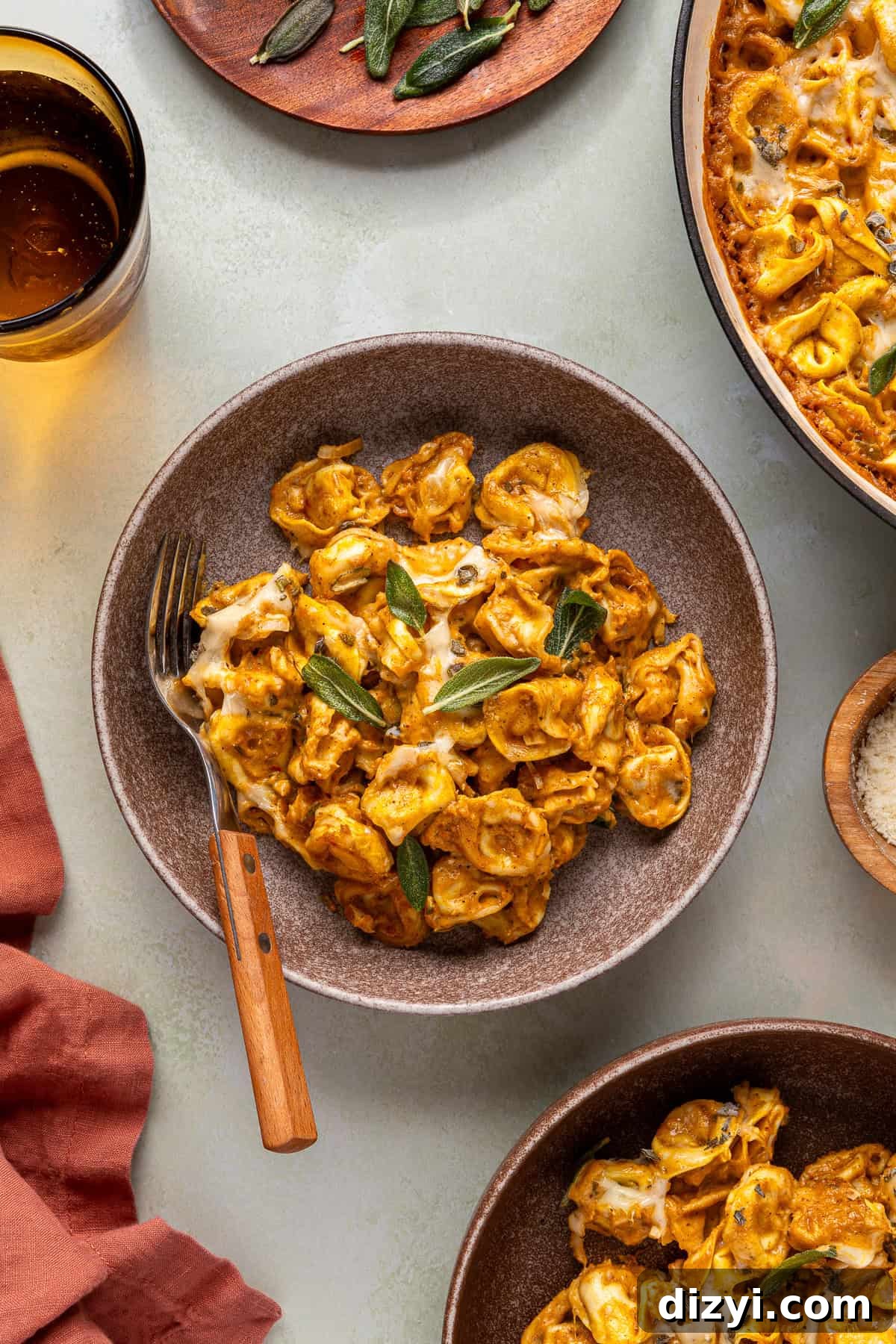 A brown dish of Creamy Pumpkin Tortellini, with a fork. Around it are a glass of water, the pan of Creamy Pumpkin Tortellini, another plate of the pasta, and a napkin.