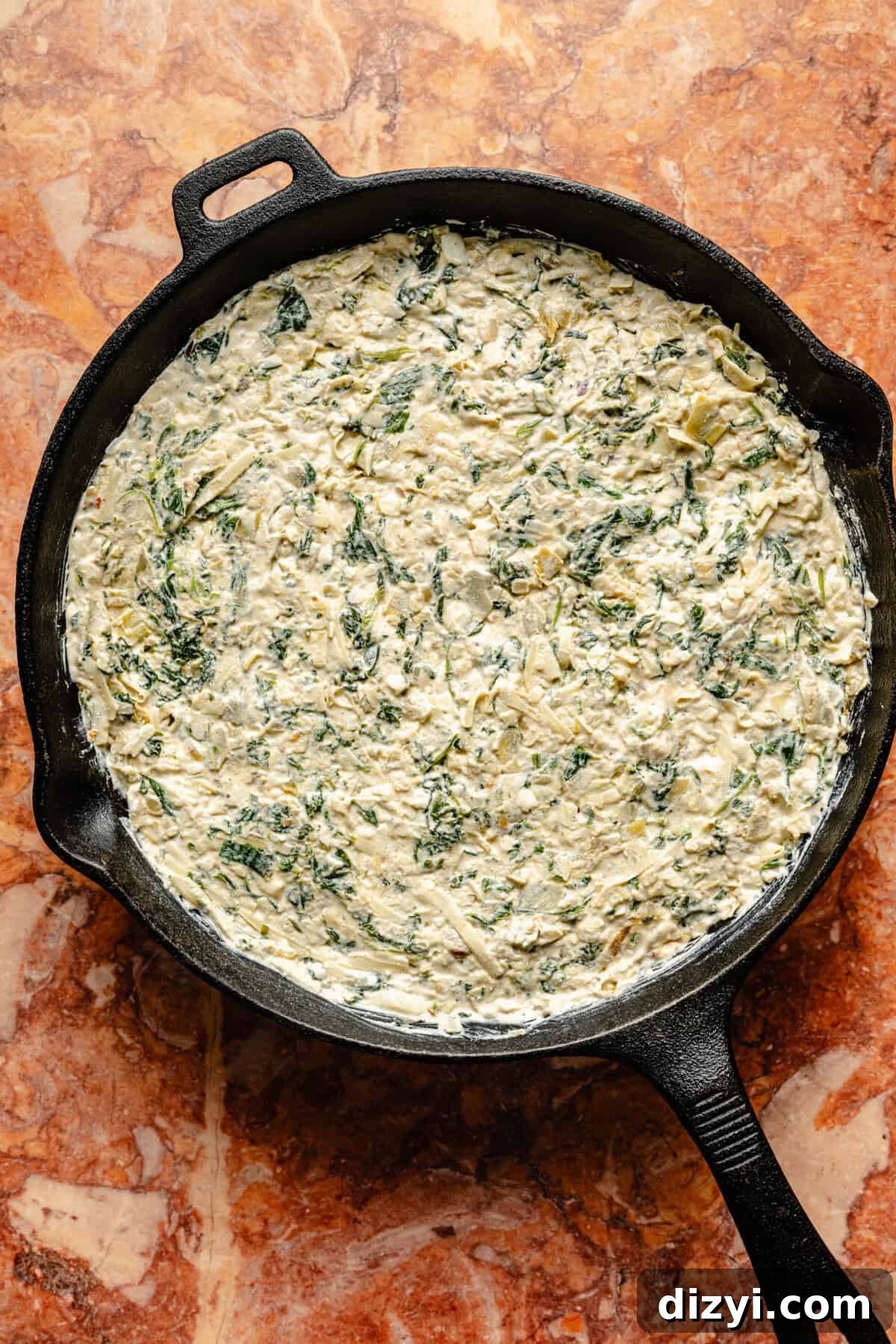 The Skillet Spinach, Artichoke, and Kale Dip in a black cast iron skillet, ready to go into the oven. The skillet is on an orange countertop with chips scattered on the counter as well.