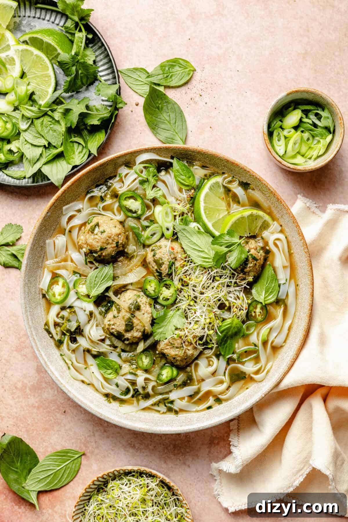 A finished bowl of Ginger-Pork Meatball Pho on a pinkish-cream countertop. A cream colored napkin is in the image, as well as dishes of green onions, broccoli sprouts, basil, cilantro, and lime wedges.