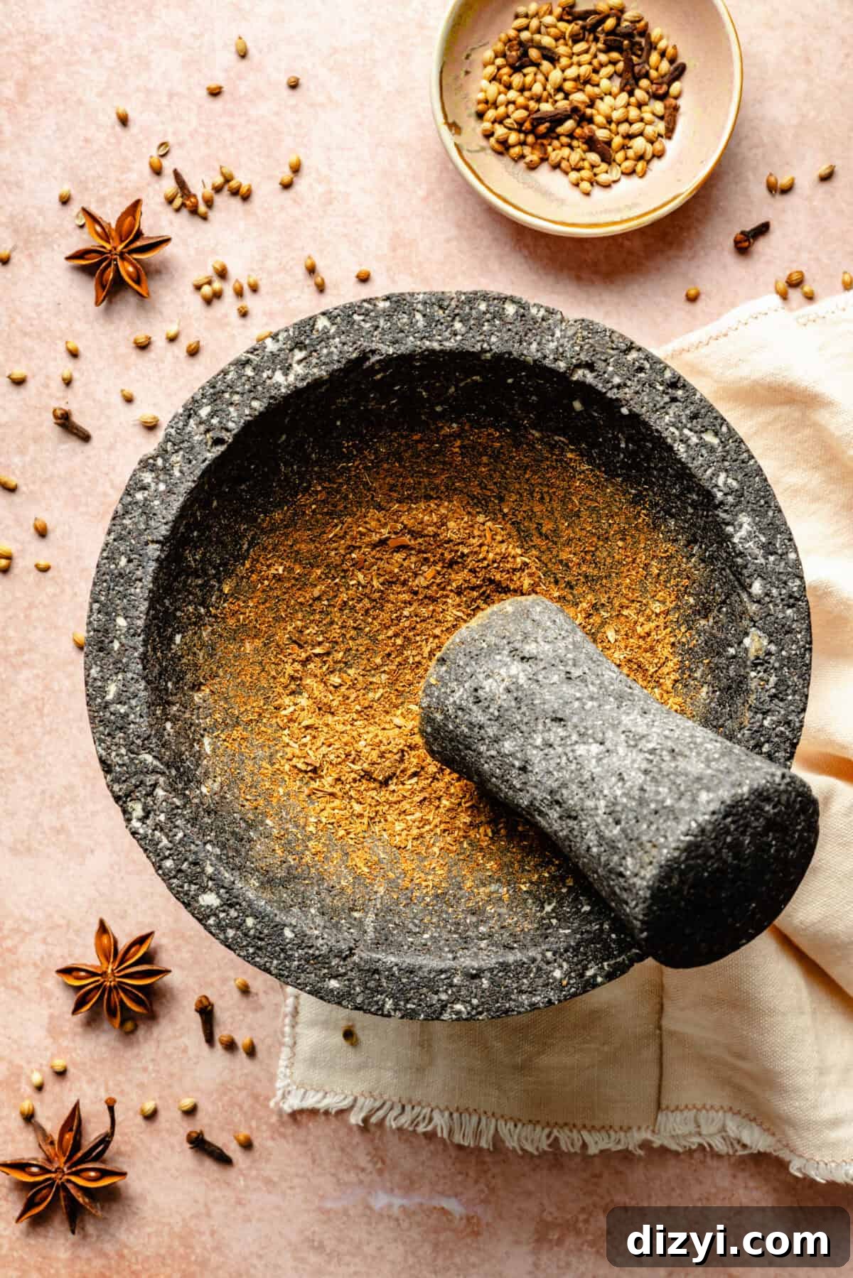 The spices needed for Ginger-Pork Meatball Pho being ground in a mortar and pestle on a pink countertop. Star anise and coriander seeds are scattered on the counter, and a small bowl of spices is at the top of the image.