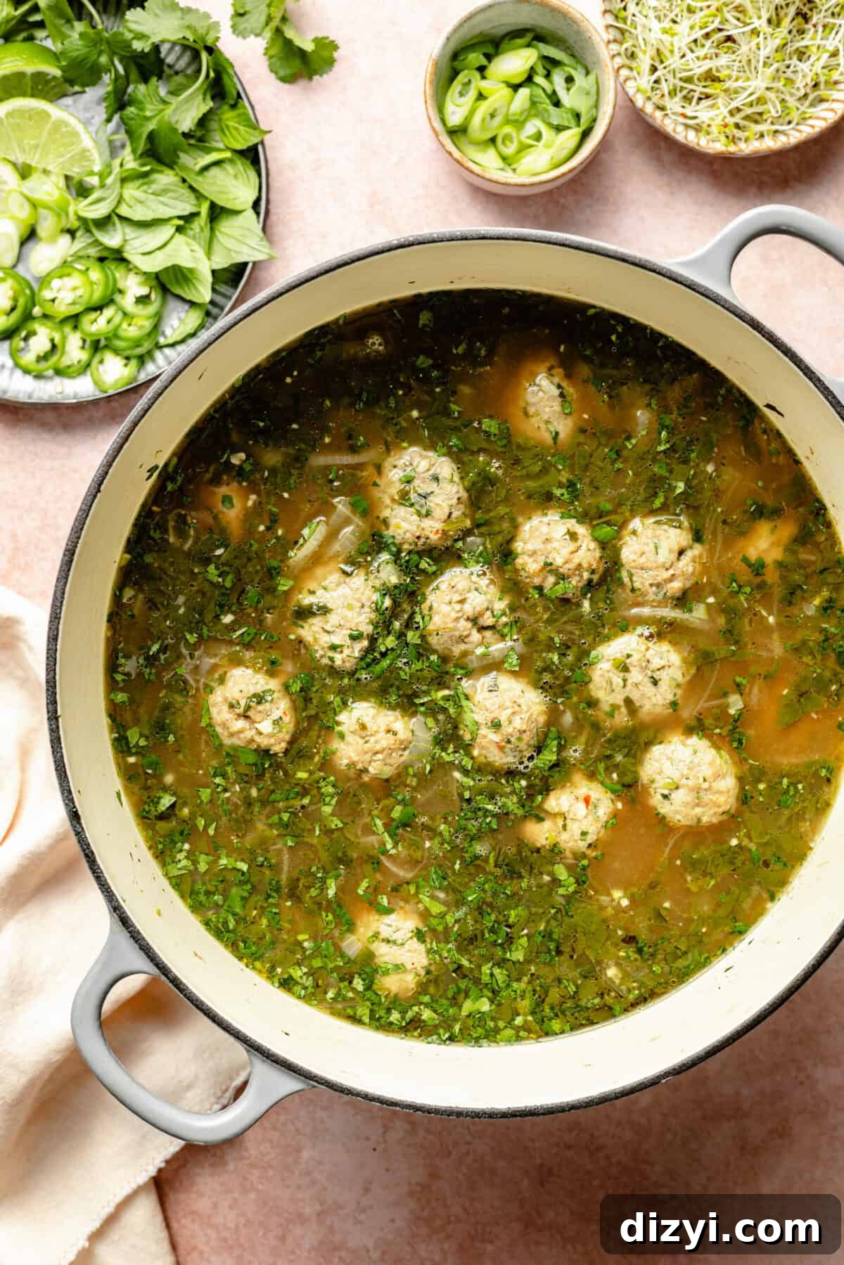 The meatballs for Ginger-Pork Meatball Pho in a stockpot with the broth and spices. A plate of jalapeño, lime wedges, and cilantro is in the top left corner, a small bowl of chopped green onions, and a small dish of broccoli sprouts are at the top of the image.