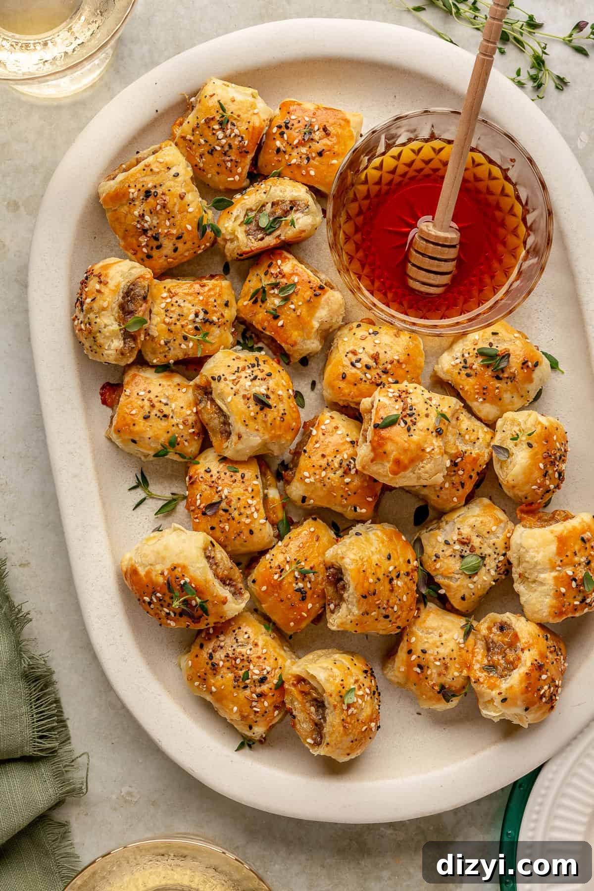 A white serving platter of Sausage Rolls, topped with thyme. A side dish of hot honey is on the dish, with a honey dipper.