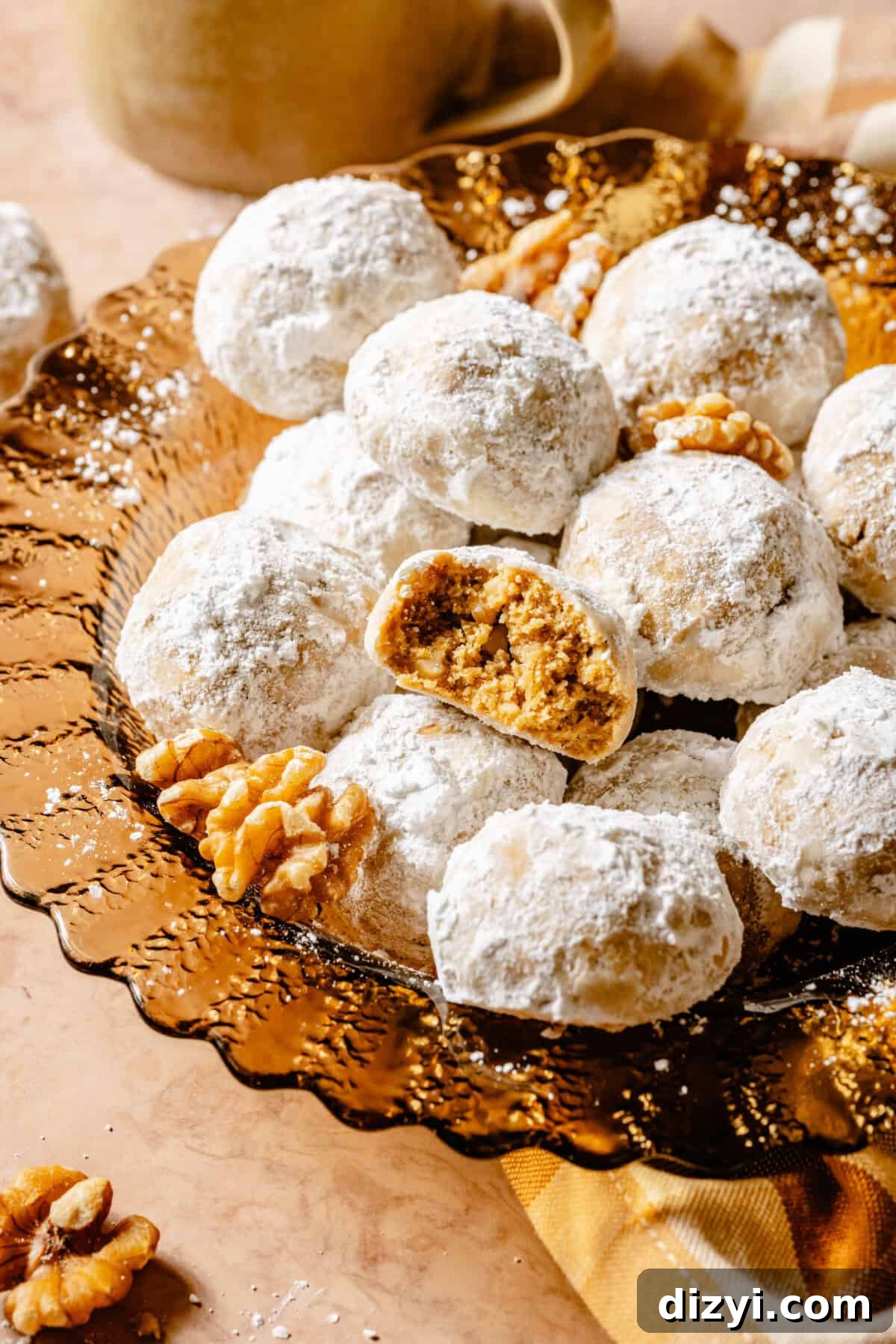 A close up shot of a finished amber plate of GoGo's Russian Teacakes on a tan countertop with a yellow checkered napkin. A few of the teacakes are on the counter, as well as a mug and a few walnuts. One teacake has a bite taken out of it.