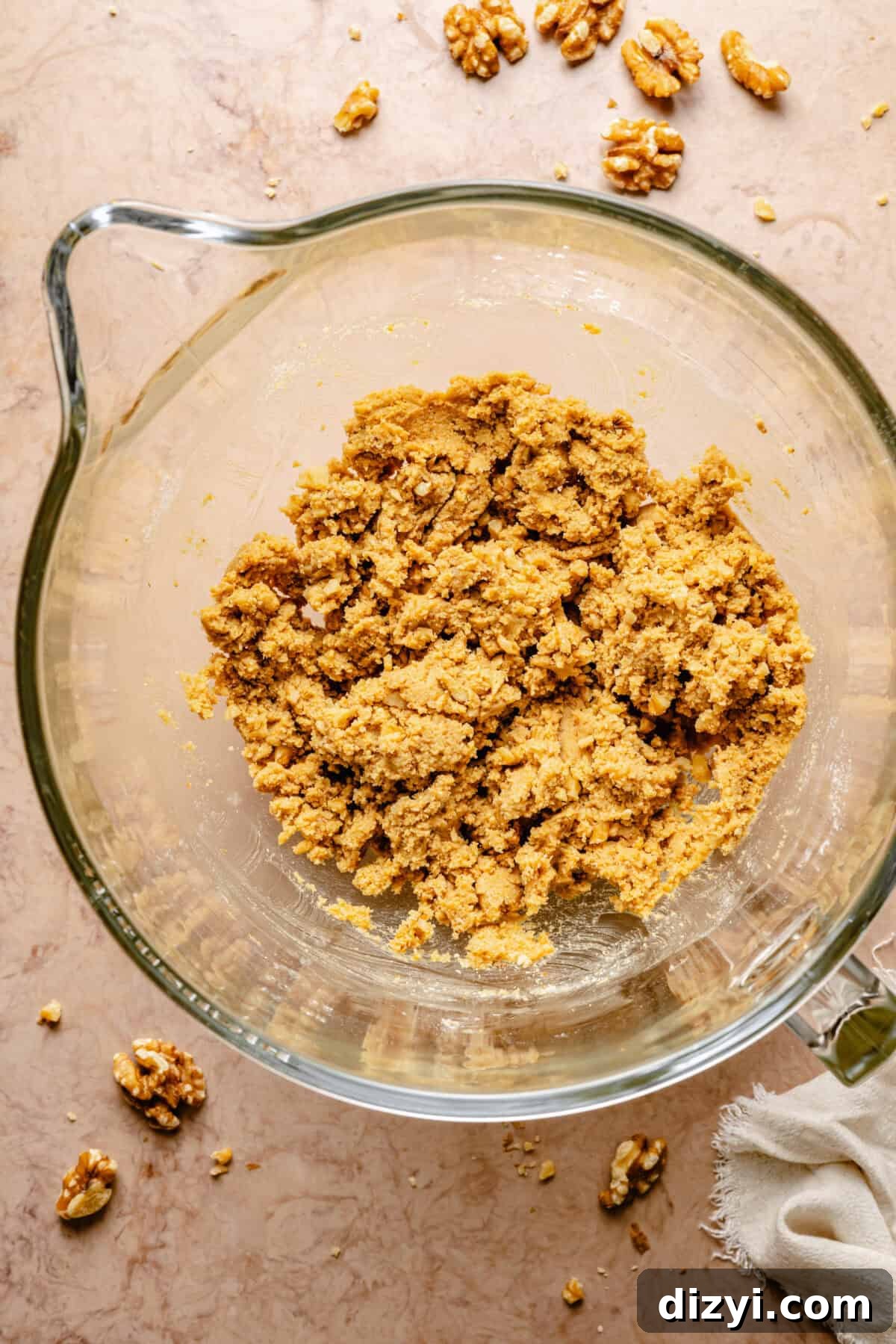 The batter for GoGo's Russian Teacakes in a clear mixing bowl on a tan countertop. Walnuts are scattered on the countertop as well.