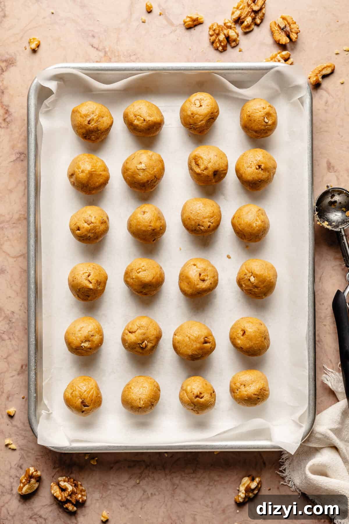 A silver sheetpan, lined with parchment paper, on a tan countertop has 24 unbaked GoGo's Russian Teacakes rolled and ready to bake.
