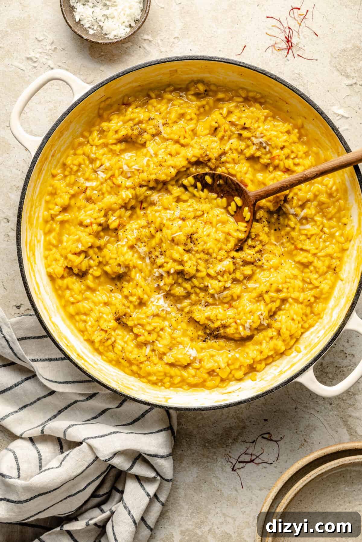Risotto Milanese in a white stockpot on a tan countertop. A wooden spoon is also in the stockpot, along with a cream and black striped napkin.