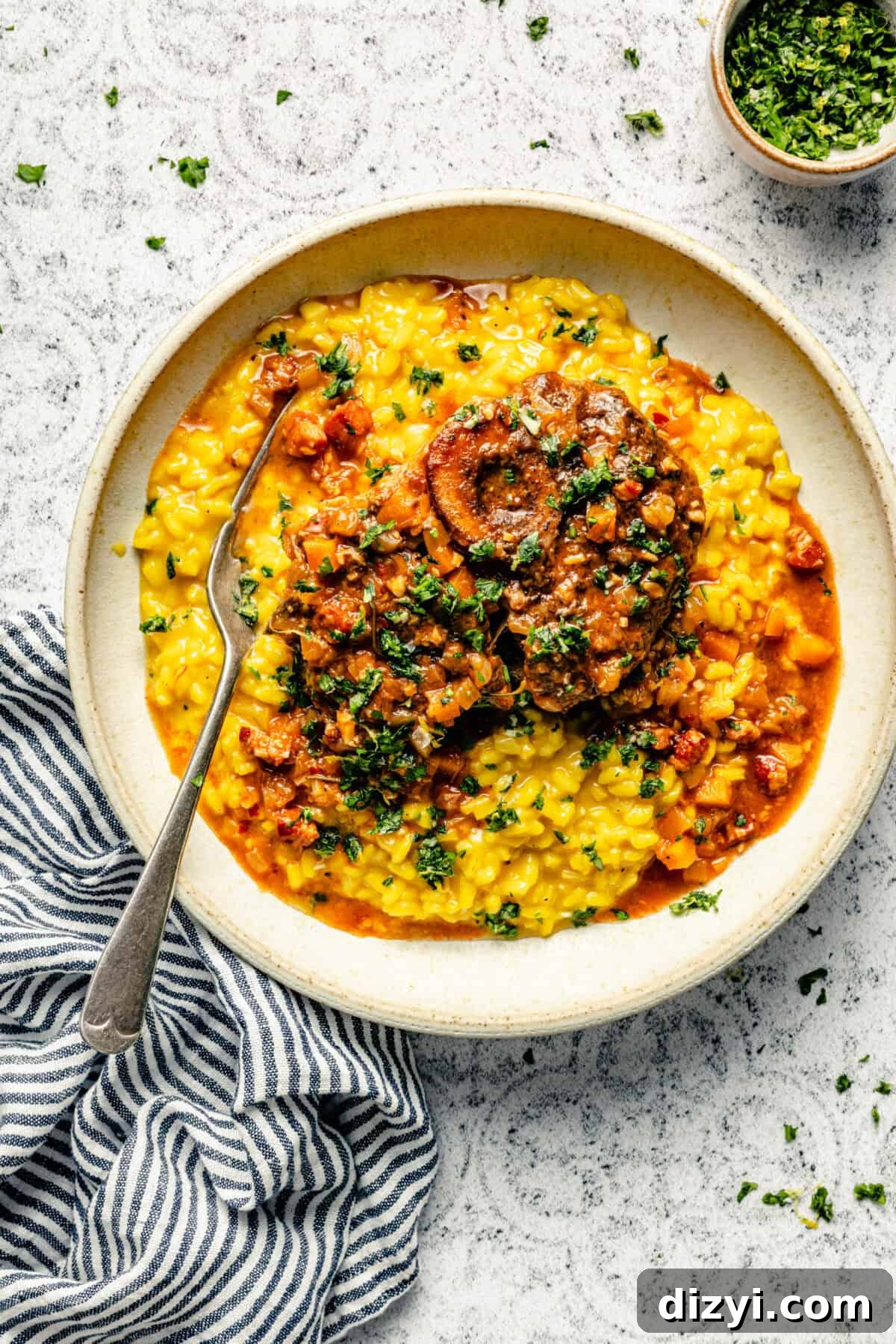 Osso Buco served atop risotto in a white dish on a black and white patterned countertop. A black and white striped napkin is also in the image.
