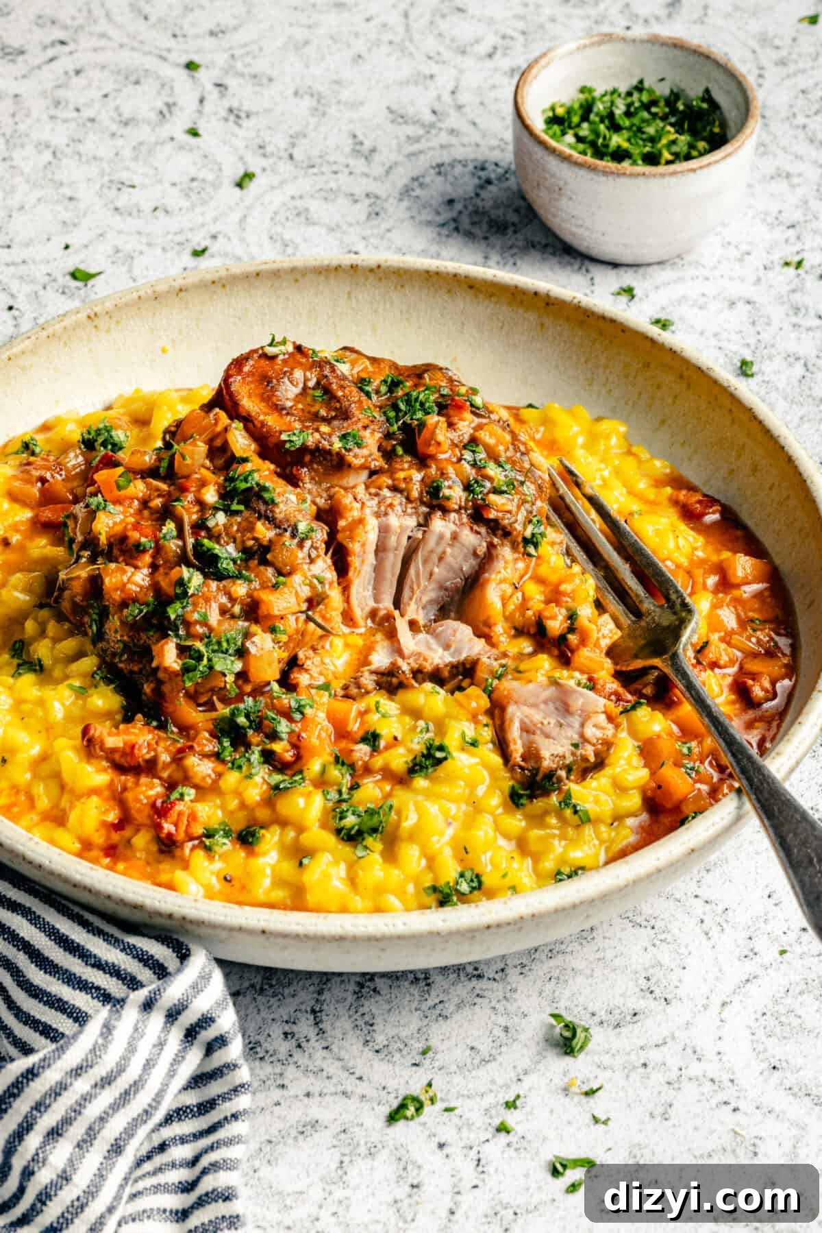 A white plate of Osso Buco served on top of risotto with a silver fork on a black and white patterned countertop. A black and white striped napkin is in the image, as well as a small bowl of chopped herbs.