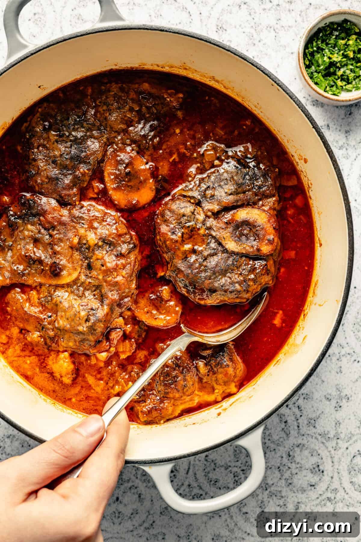 Osso Buco being served with a silver spoon from a stockpot on a black and white patterned countertop.