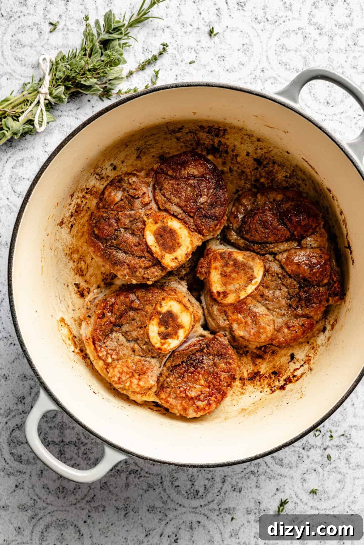 A stock pot with three large veal shanks browning in the bottom of it. A bundle of rosemary, thyme, and oregano is tied together with kitchen twine in the top of the image. The stock pot sits on a white and black patterned countertop.