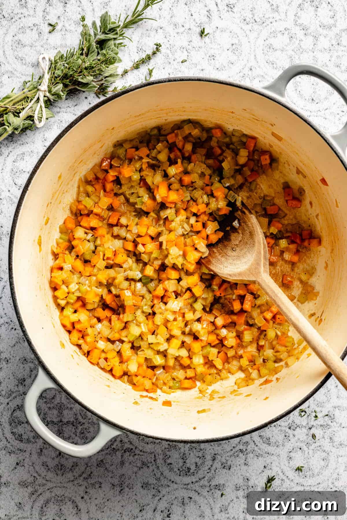Diced onion, carrot, and celery cooking in a stockpot on a white and black patterned countertop. A wooden spoon is in the pot, and a bundle of herbs is tied with kitchen twine at the top of the image.