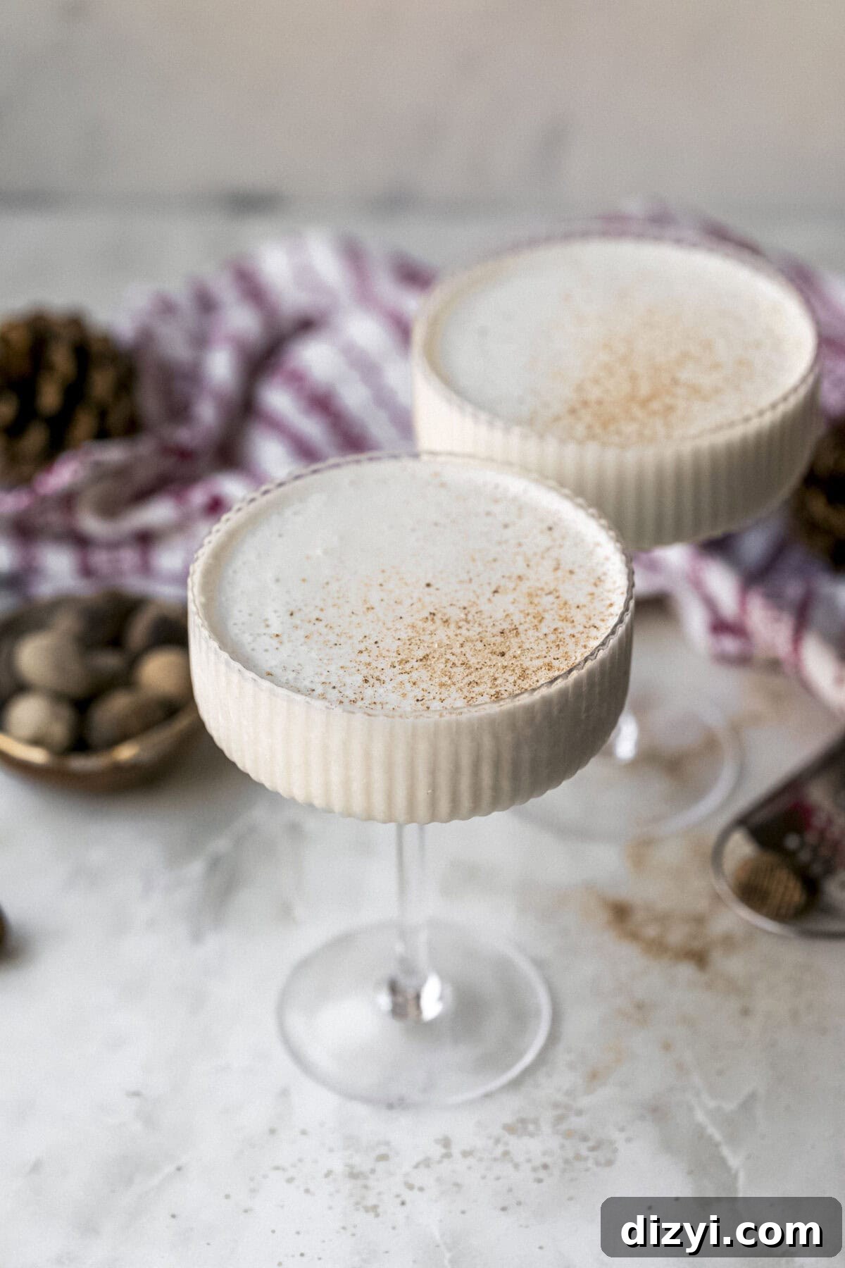 Two finished coupe glasses of Brandy Freeze Cocktail on a marble countertop. Nutmeg and pinecones are on the counter, along with a red and white napkin.