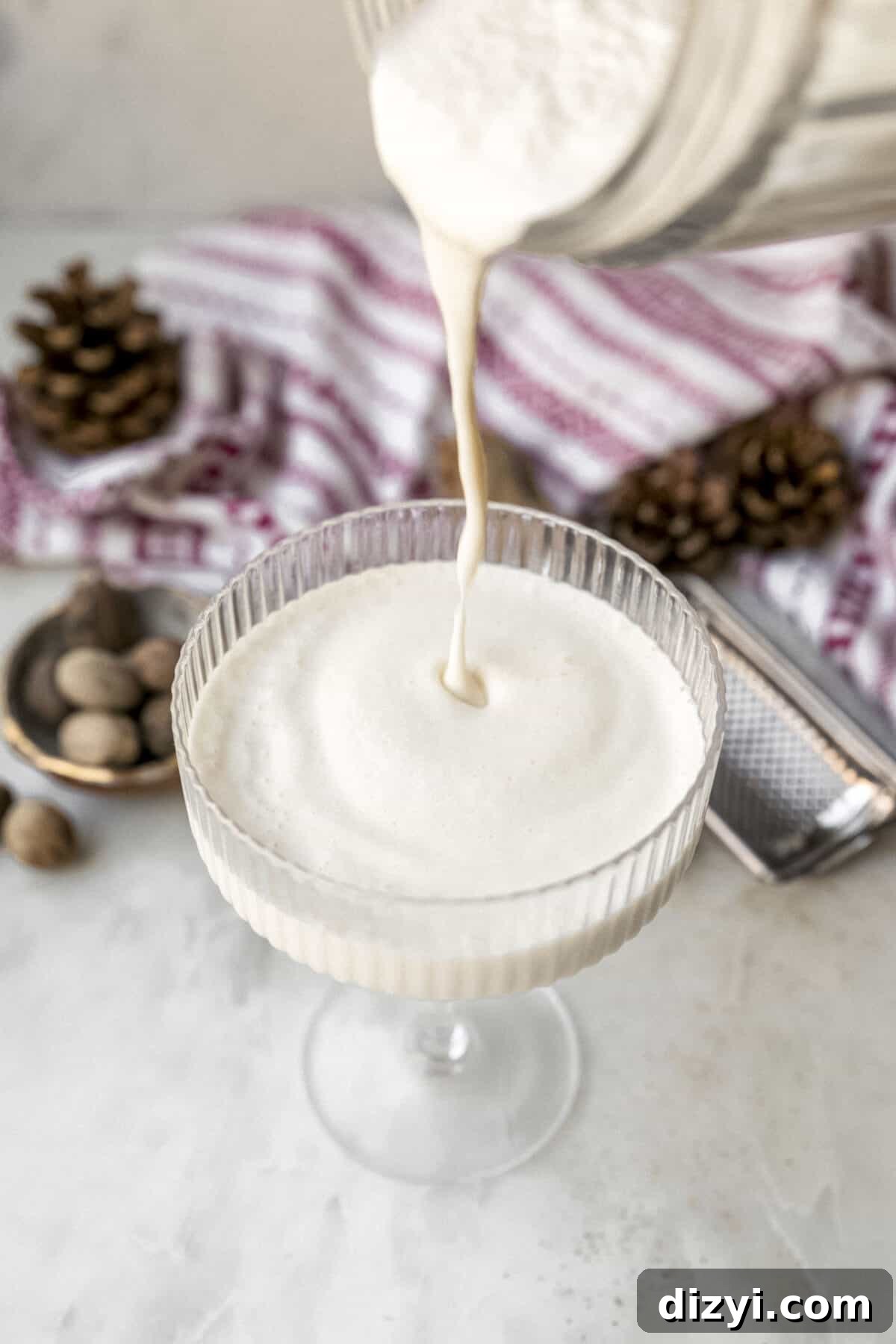 The blended Brandy Freeze Cocktail being poured into a couple glass on a marble countertop. Nutmeg, pinecones, and red and white napkins are visible on the countertop as well.