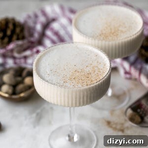 Two finished coupe glasses of Brandy Freeze Cocktail on a marble countertop. Nutmeg and pinecones are on the counter, along with a red and white napkin.