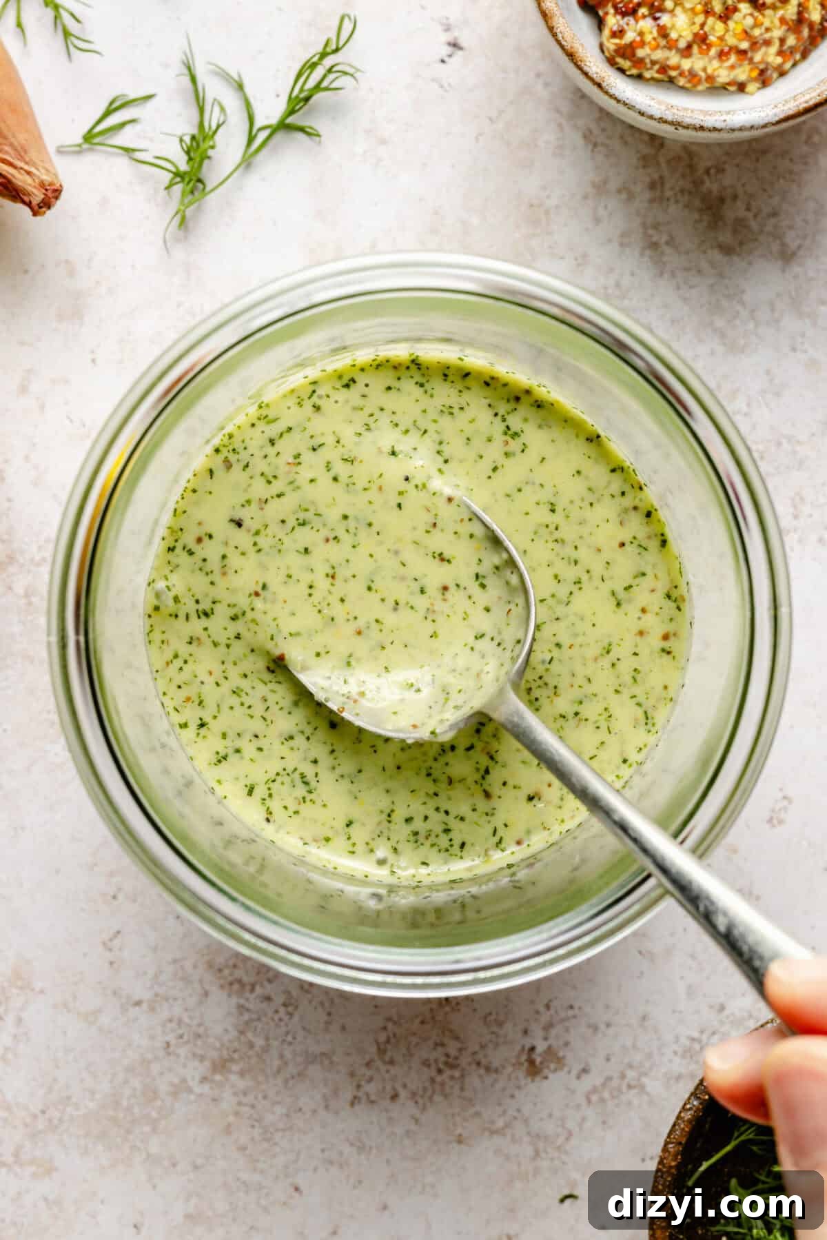 A clear jar of shallot-dijon vinaigrette for the Moroccan-Inspired Roasted Chicken Salad with a silver spoon dipped into it.