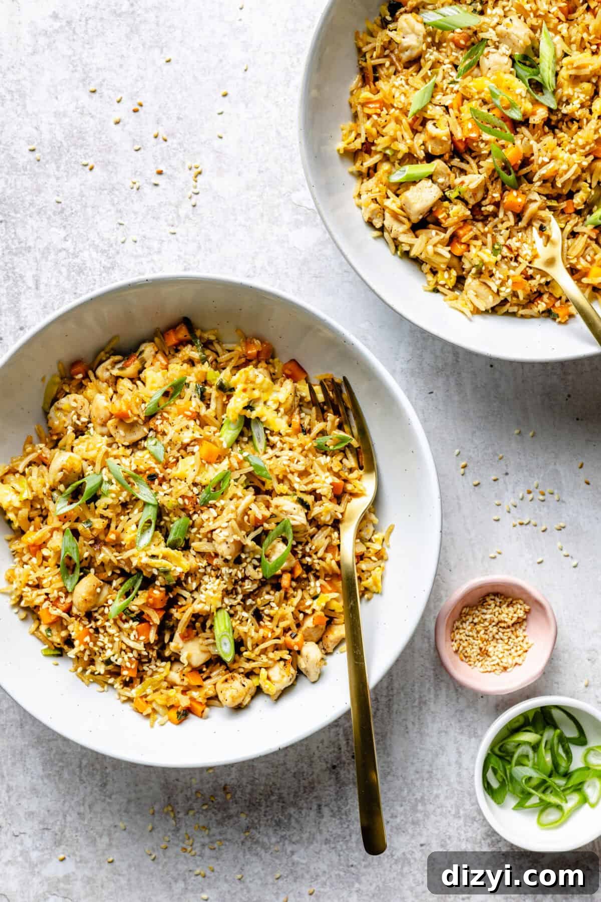 Two finished white bowls of Simple Chicken Fried Rice on a gray countertop. A small pink dish of toasted sesame seeds and a small white dish of sliced green onions are also on the countertop.