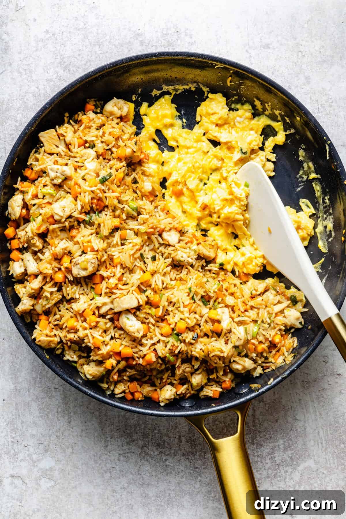 A black skillet with a gold handle is on a gray countertop with rice, chicken, and veggies, as well as a couple of eggs being scrambled on the side. A white spatula is in the skillet.