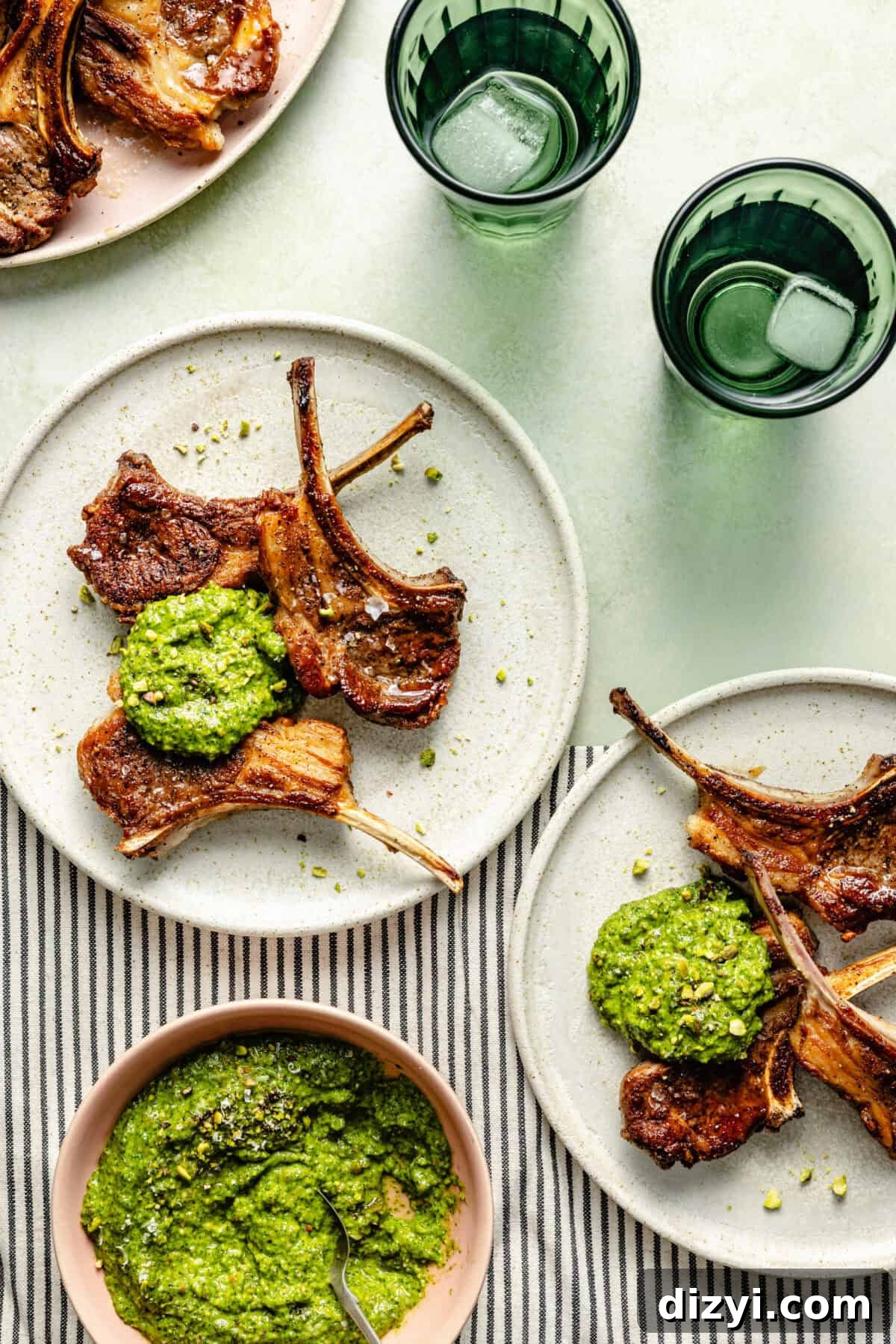 White plates with grilled lamb chops and a dollop of arugula-pistachio pesto on a green and striped background