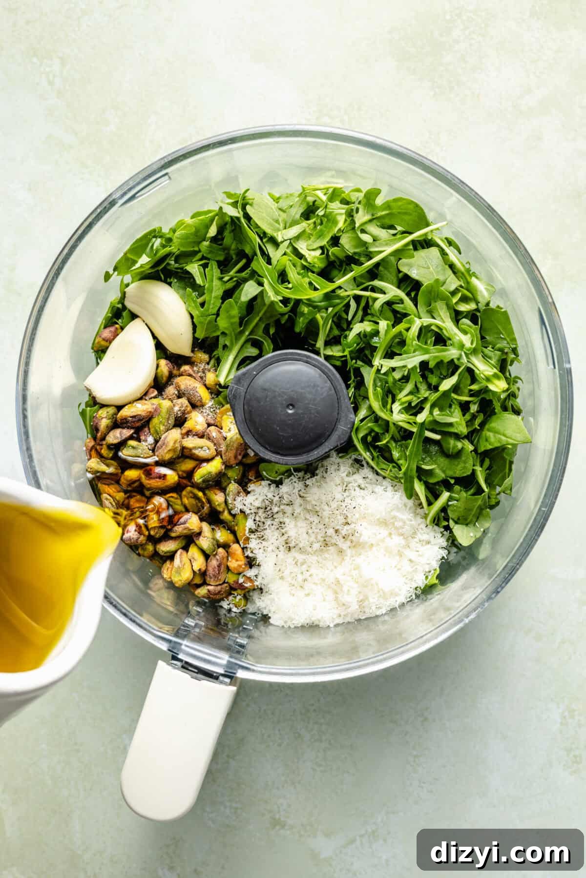 a bowl of a food processor filled with arugula, pistachios, garlic, and parmesan cheese