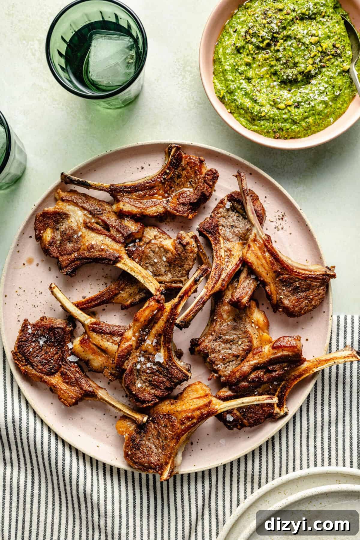 a light pink plate filled with grilled lamb chops and a side bowl of arugula-pistachio pesto on a green background