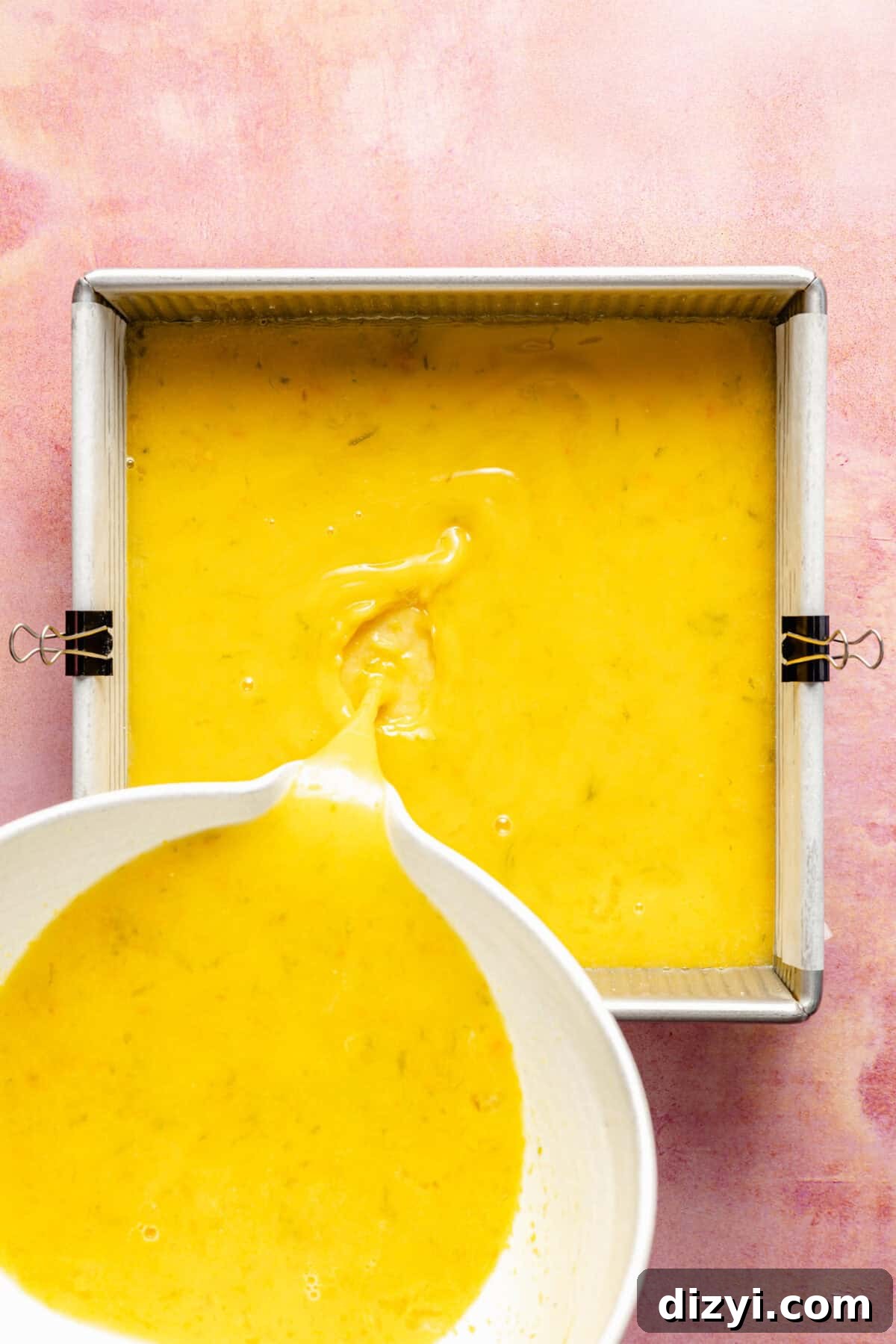 a bowl of citrus filling being poured into a square lined baking tray on a pink background