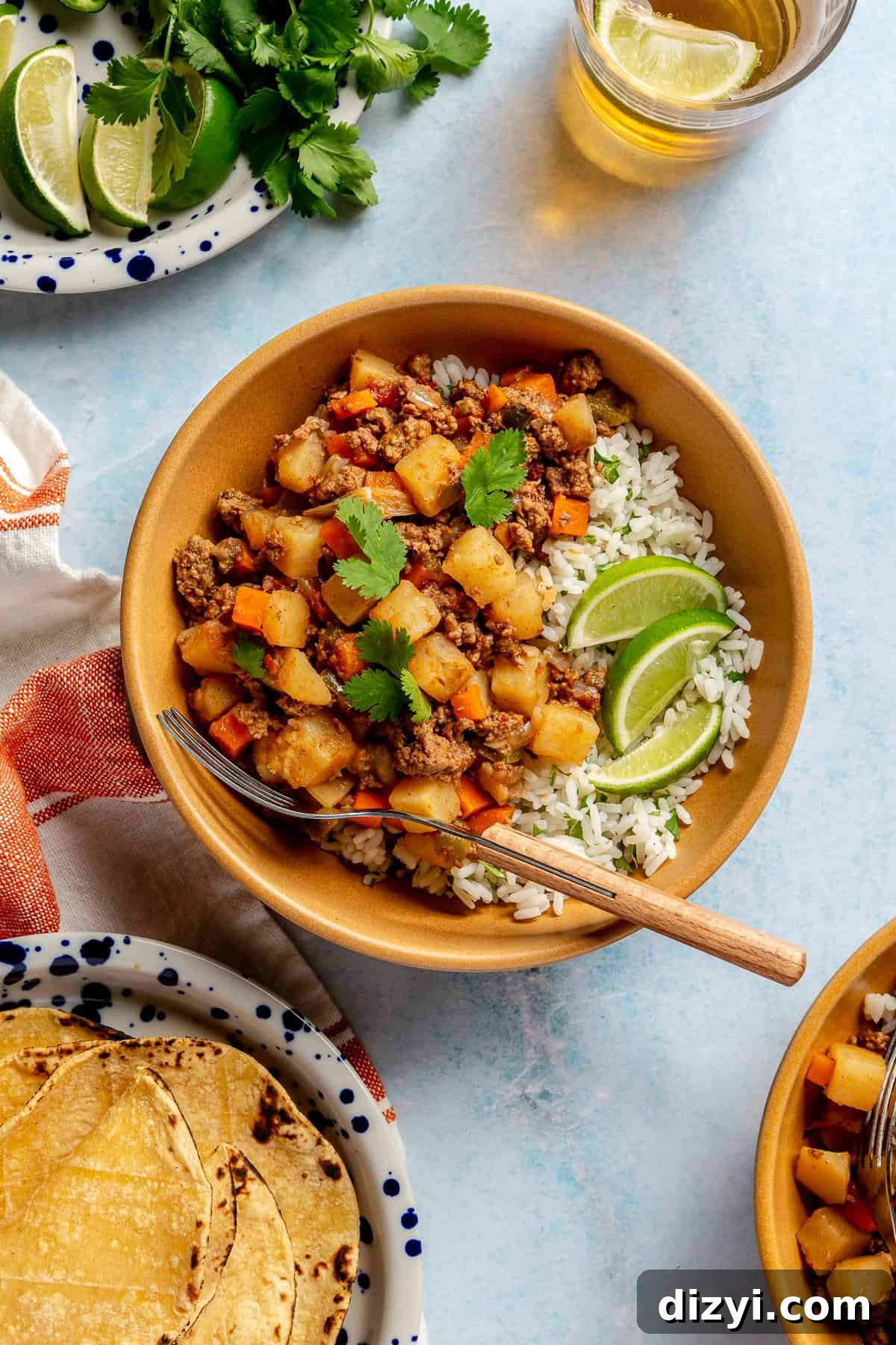 A finished bowl of Crockpot Mexican Picadillo in an orange bowl with a wooden fork. The picadillo is served over a bed of rice with three lime slices and some sprigs of cilantro as garnishes.