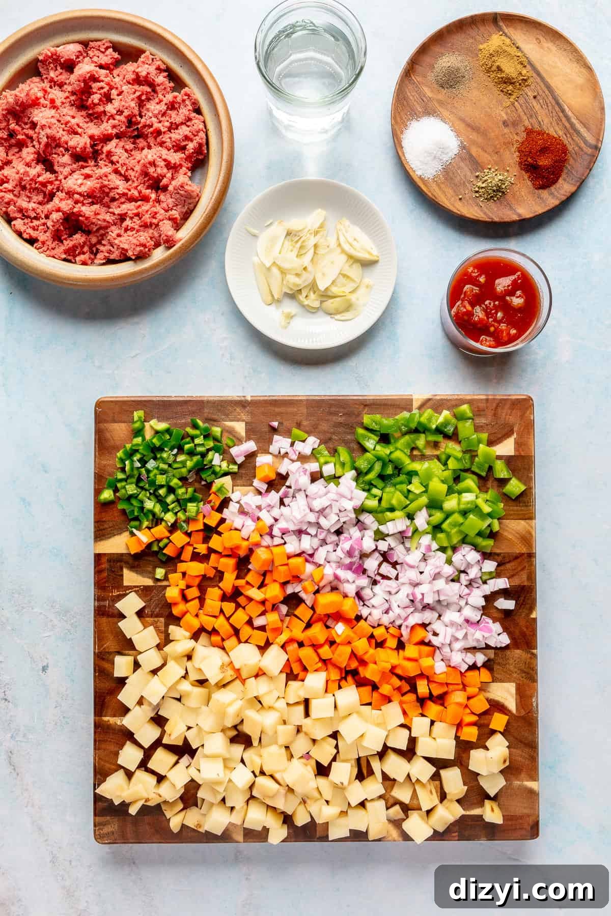 The ingredients for the Crockpot Mexican Picadillo chopped and on a wooden cutting board. A bowl of ground beef is in the top left, a dish of spices in the top right, as well as a dish of sliced garlic, a glass of water and a container of crushed tomatoes.