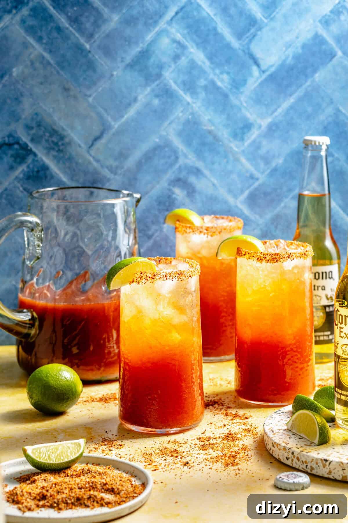three glasses of Michelada mix with a bottle of cold beer and limes on a blue background