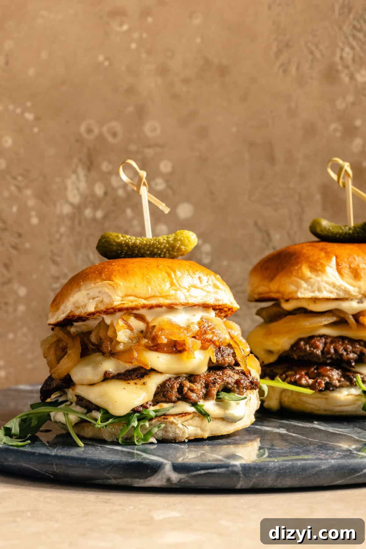 two smashburgers with cheese, caramelized onions, and arugula with a cornichon garnish on blue marble plates