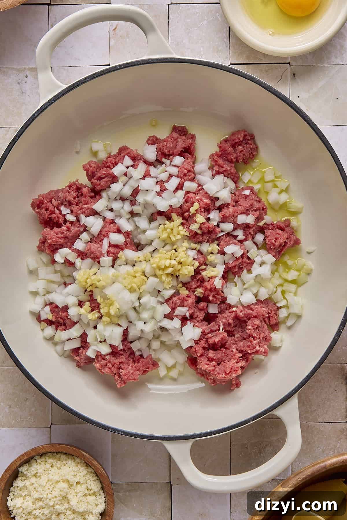 a white cast-iron braiser with raw onions, garlic, and ground beef