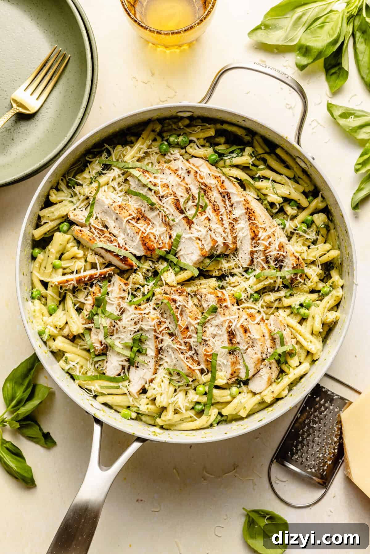 A skillet with Creamy Pesto Chicken Pasta on a white countertop. There is also a cheese grater and a block of cheese visible, as well as a few basil leaves.