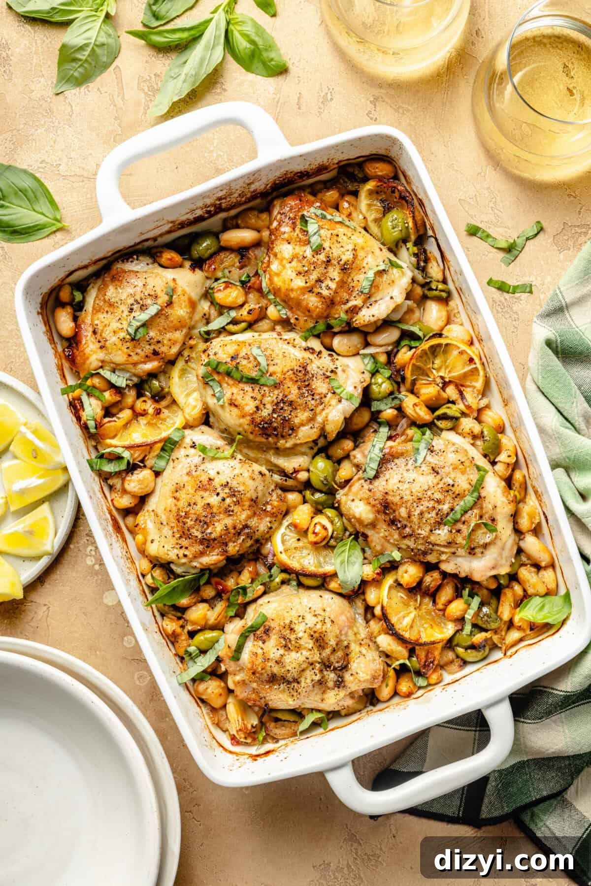 a white baking dish with baked chicken thighs, beans, lemons, and herbs