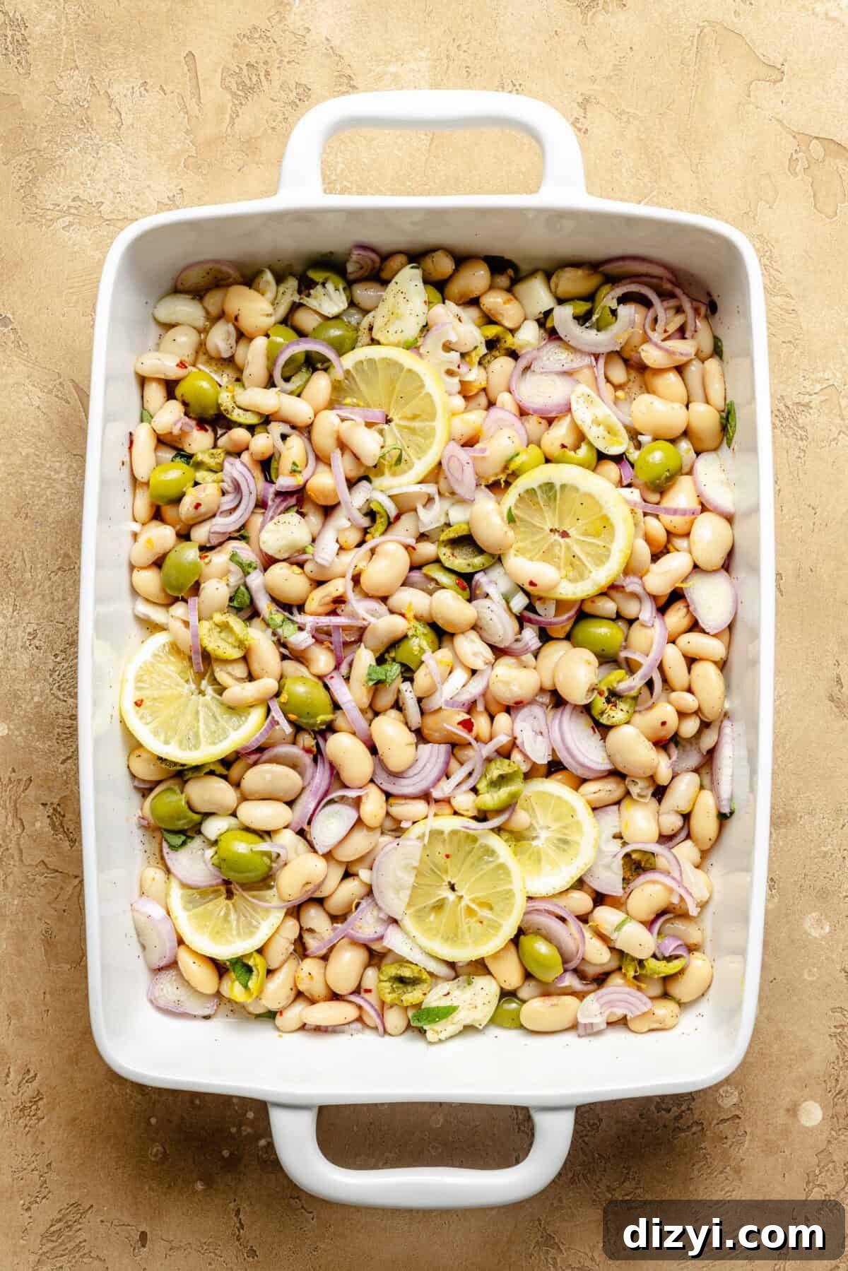 a white baking dish with white beans, olives, red onions, and sliced lemons on a tan background