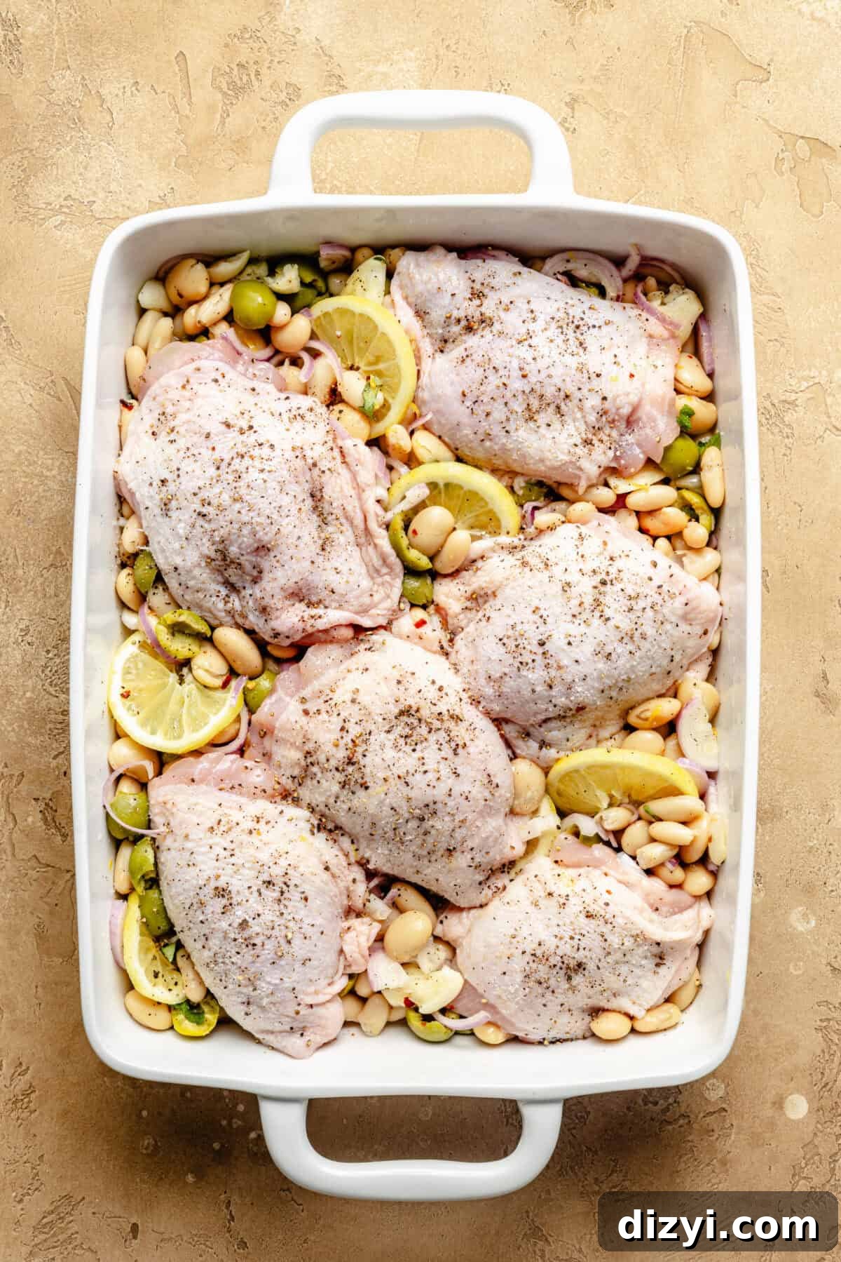 a white baking dish on a tan background filled with white beans, olives, lemons, and seasoned chicken thighs