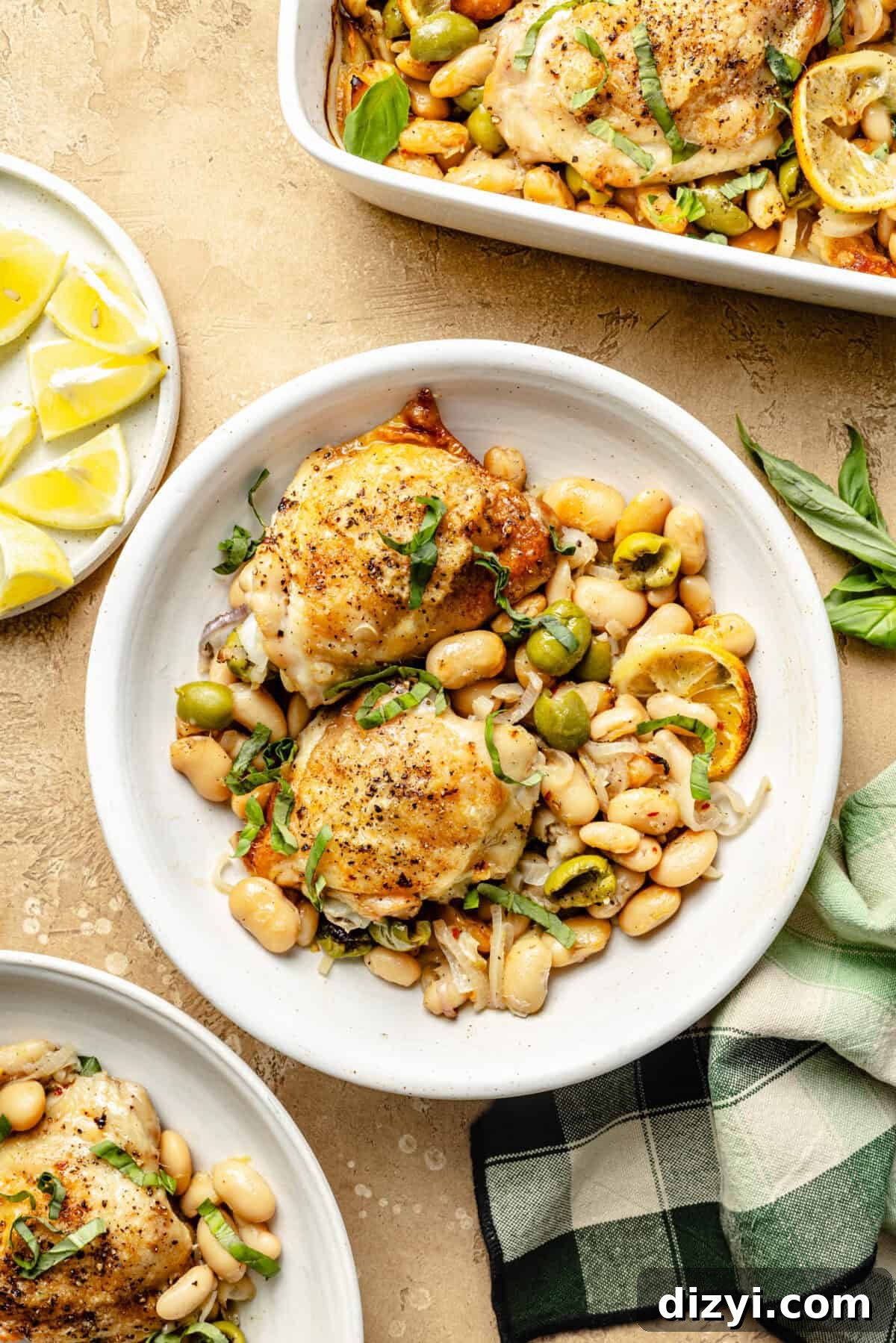 a white shallow bowl with seared chicken thighs, white beans, olives, lemons, and fresh basil