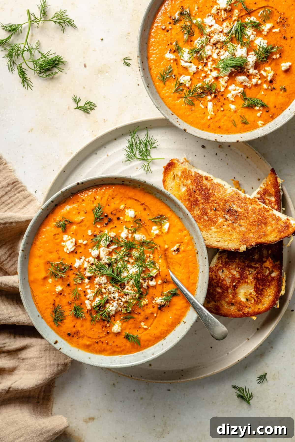 two bowls of tomato soup garnished with feta cheese and dill and served alongside a grilled cheese sandwich