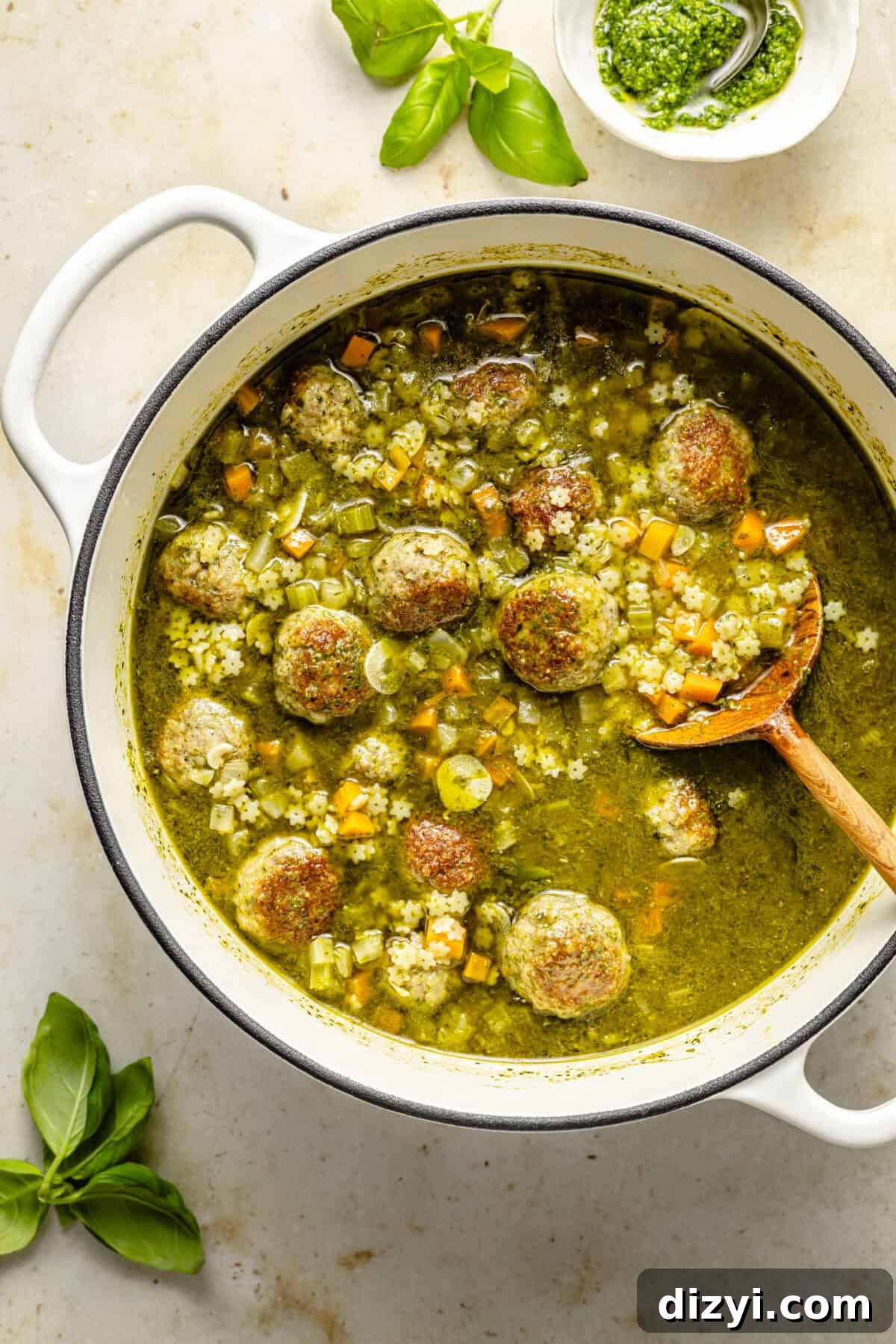 a large white pot with pesto meatball soup and pasta; a garnish of fresh basil leaves and basil pesto are on the side