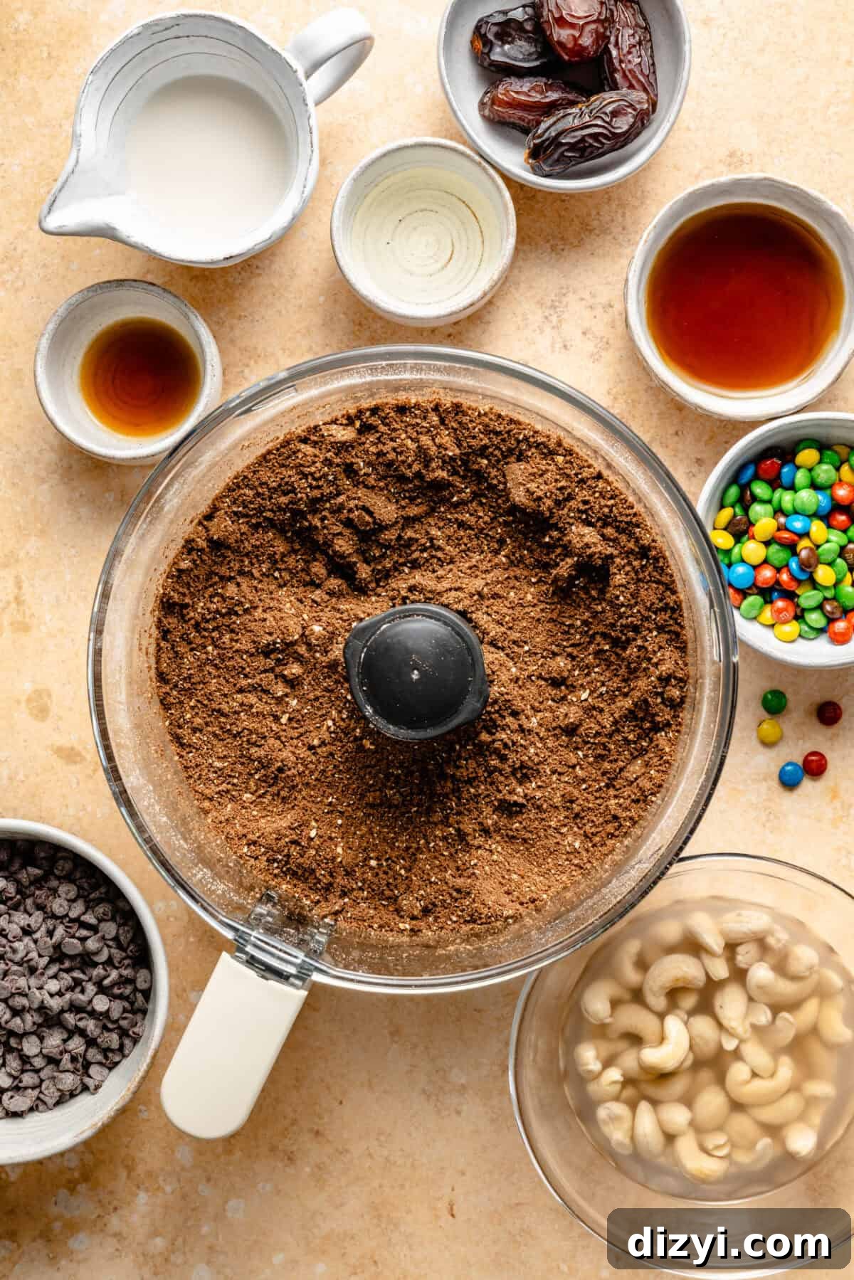 Cosmic Brownie Energy Bites Recipe - No-Bake Chocolate Snack 3 a food processor with crushed cocoa and cookie crumbs, plus small bowls of cashews, mini M&Ms, chocolate chips, and maple syrup