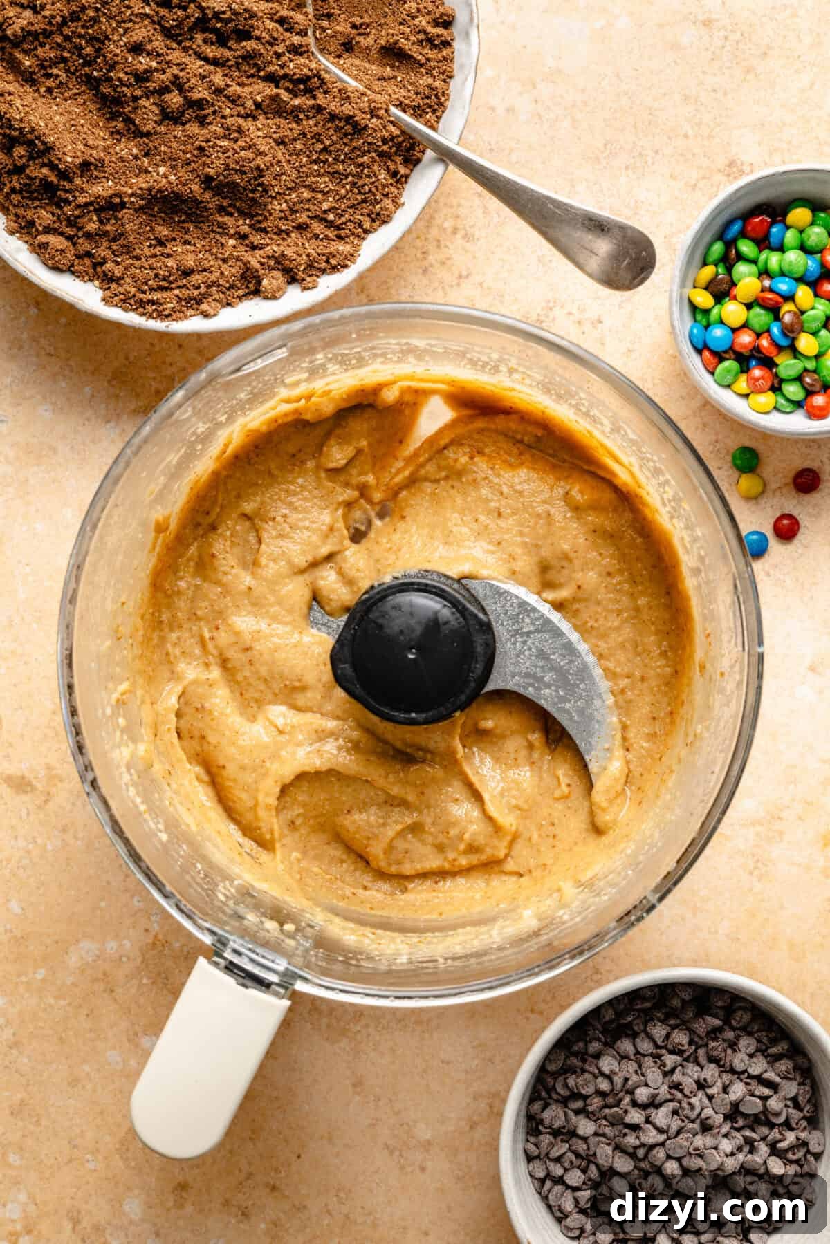 Cosmic Brownie Energy Bites Recipe - No-Bake Chocolate Snack 4 a food processor with a creamy nut butter mixture and two small bowls of mini chocolate chips and mini M&Ms